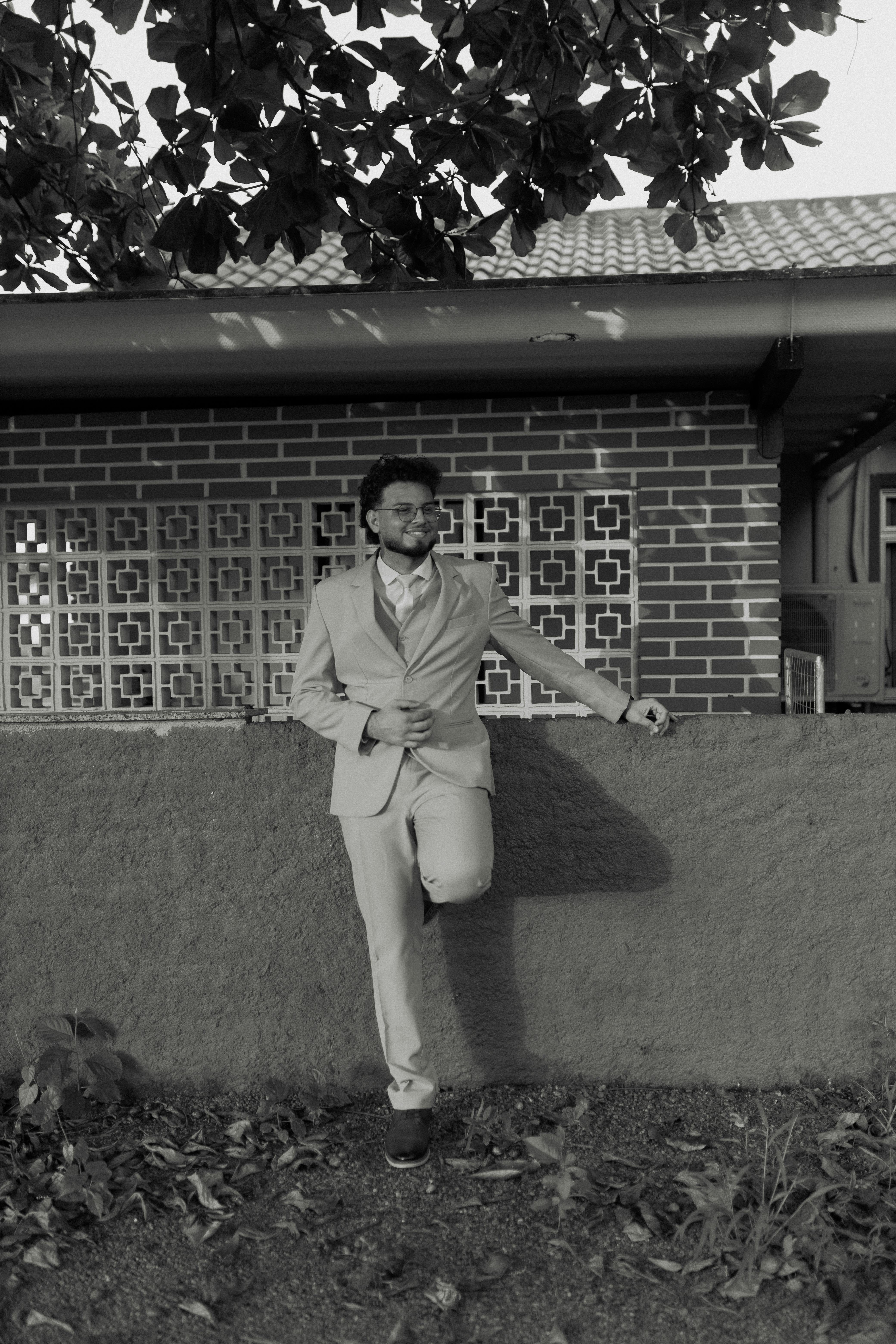 Gratuit Portret alb-negru al unui bărbat în costum, rezemat de un zid de cărămidă, în aer liber, sub un copac. Fotografie de stoc