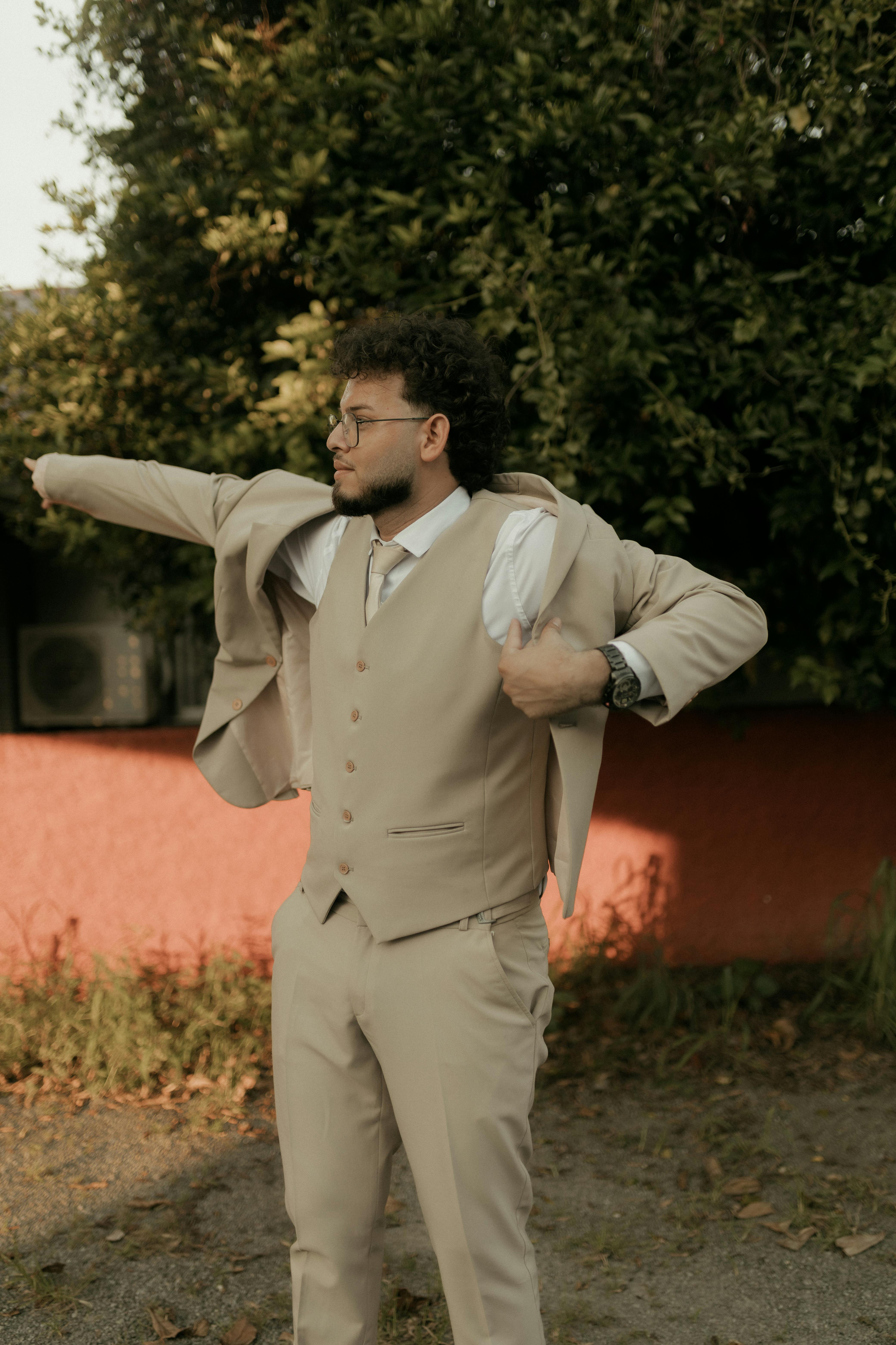 Free Young man in a stylish beige suit posing outdoors near a red wall during the day. Stock Photo