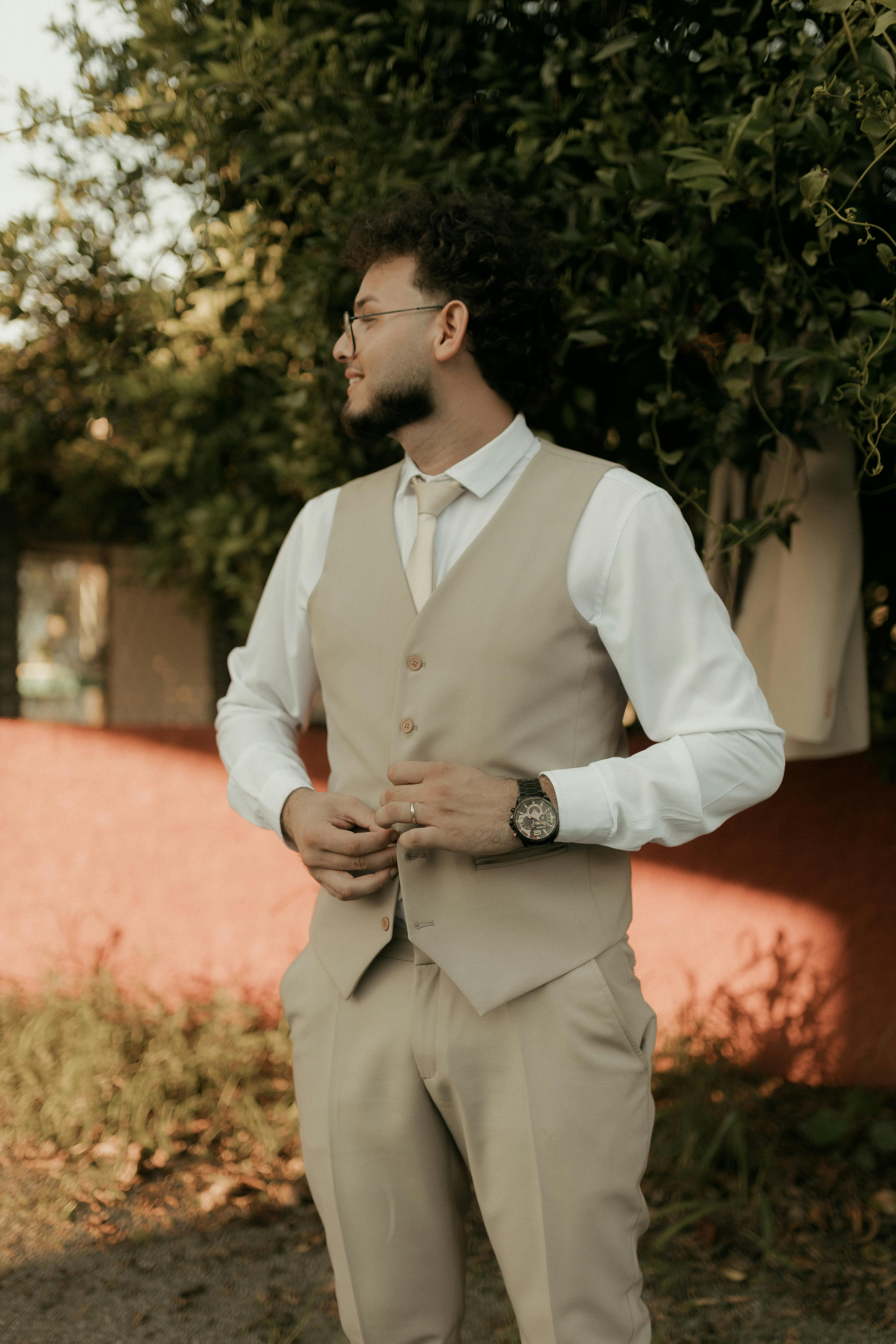 Free Elegant young man in beige suit and glasses standing outdoors in a garden setting. Stock Photo