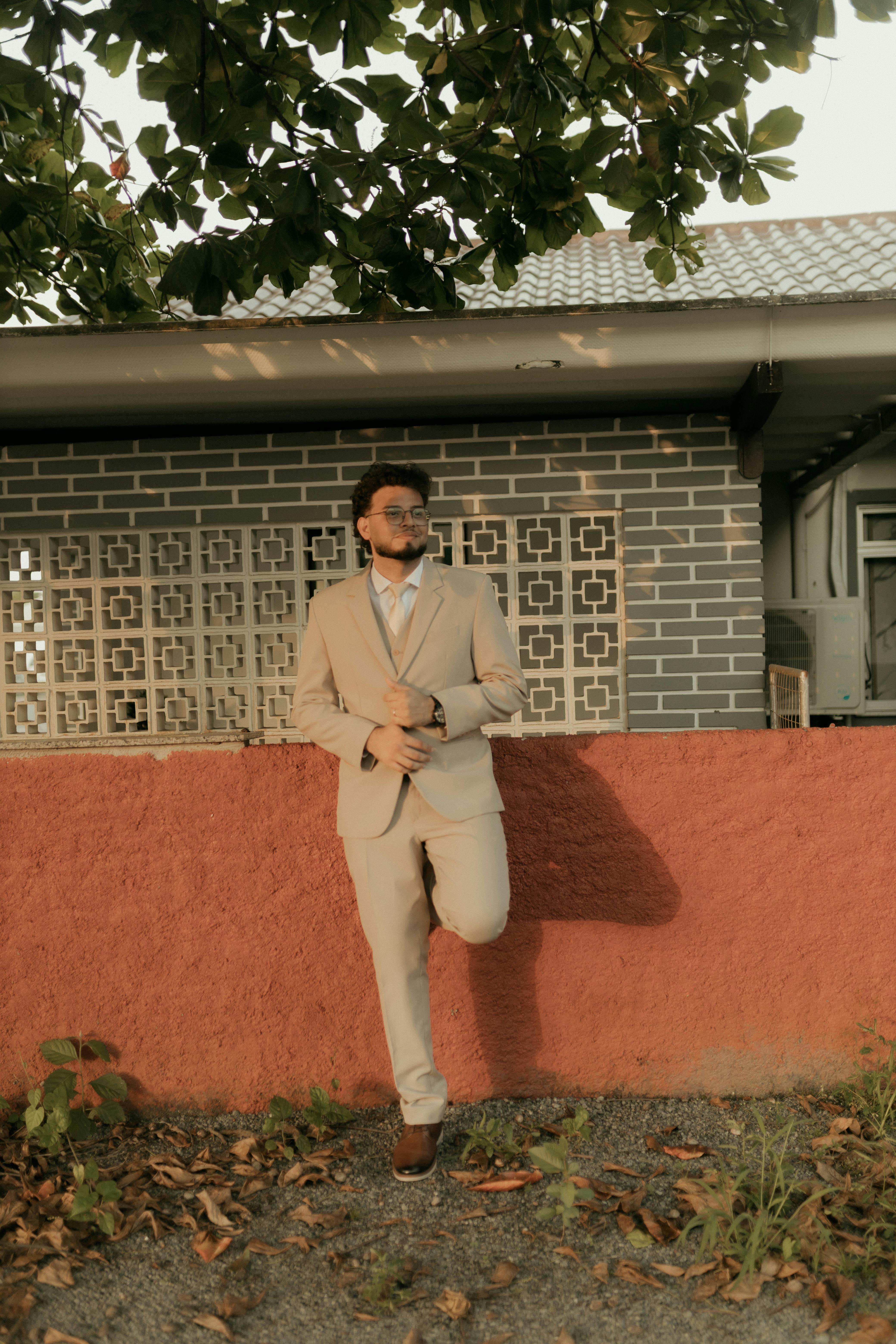 Δωρεάν Εκλεπτυσμένος άντρας με μπεζ κοστούμι ακουμπισμένος σε έναν τοίχο κάτω από ένα δέντρο έξω από ένα κομψό κτίριο. Φωτογραφία από στοκ φωτογραφιών