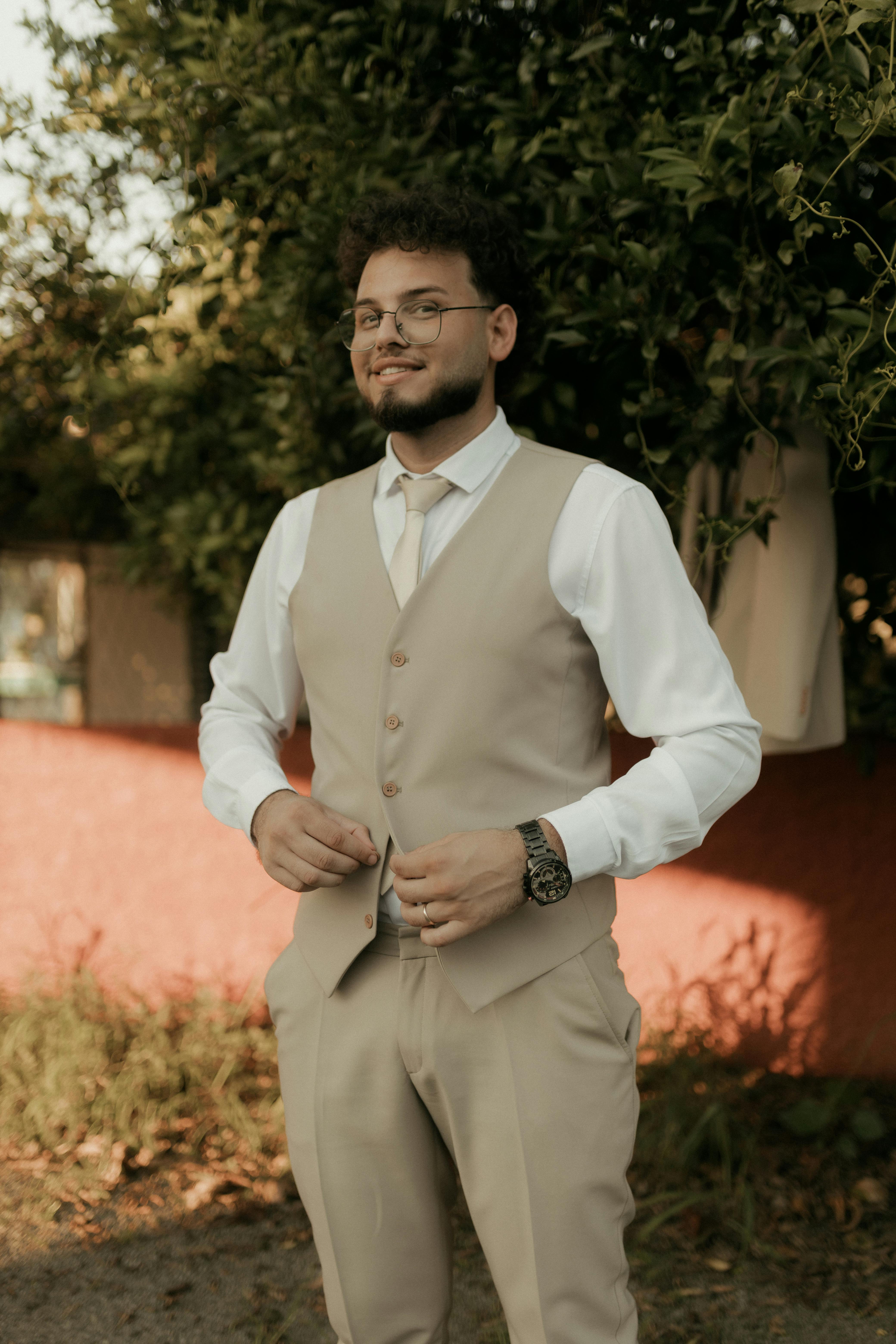 Gratis Ung mann i beige dress og slips, står utendørs med et smil i varmt sollys. Arkivbilde