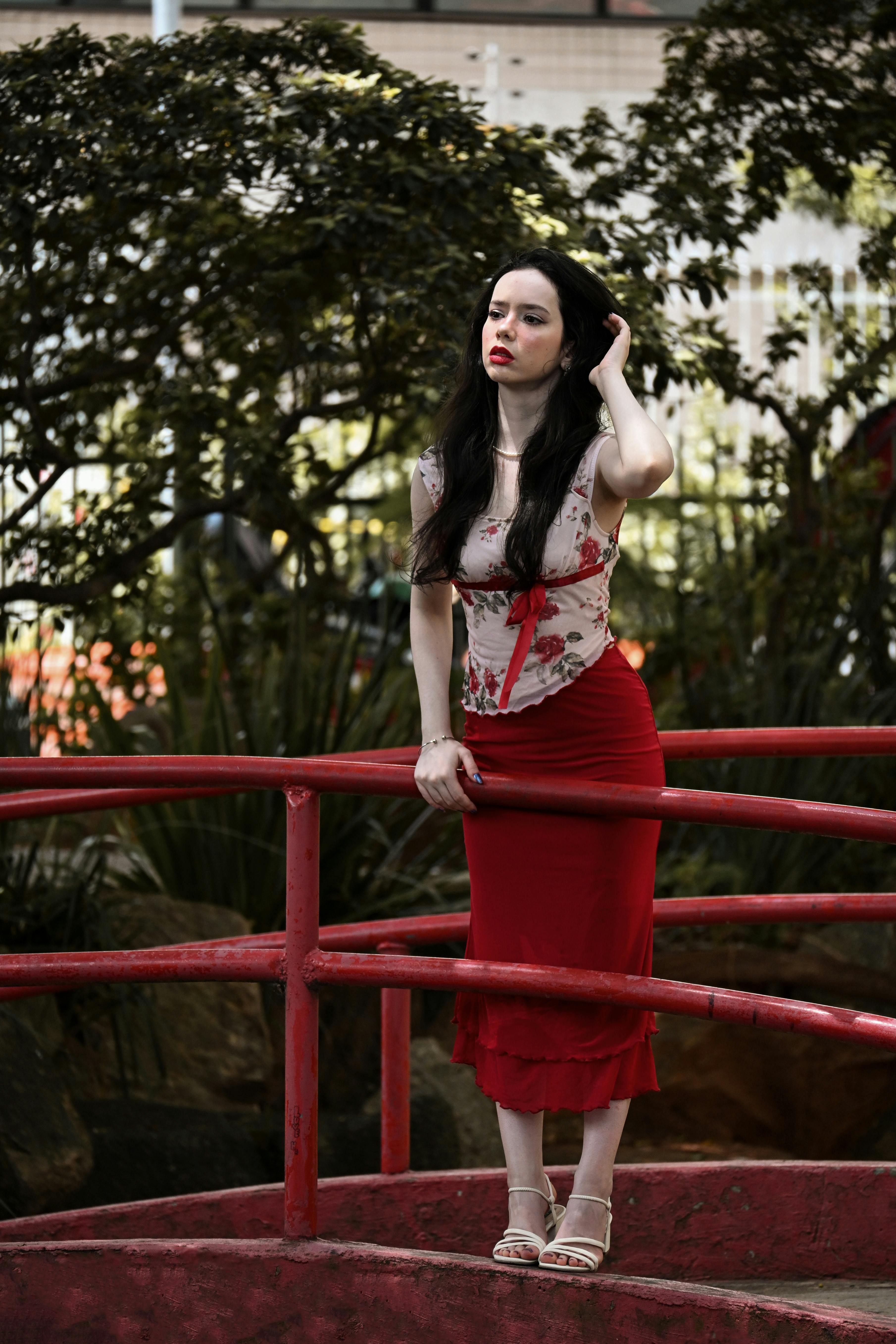 Miễn phí Một người phụ nữ mặc váy đỏ đứng duyên dáng bên lan can công viên, xung quanh là cây xanh. Ảnh lưu trữ