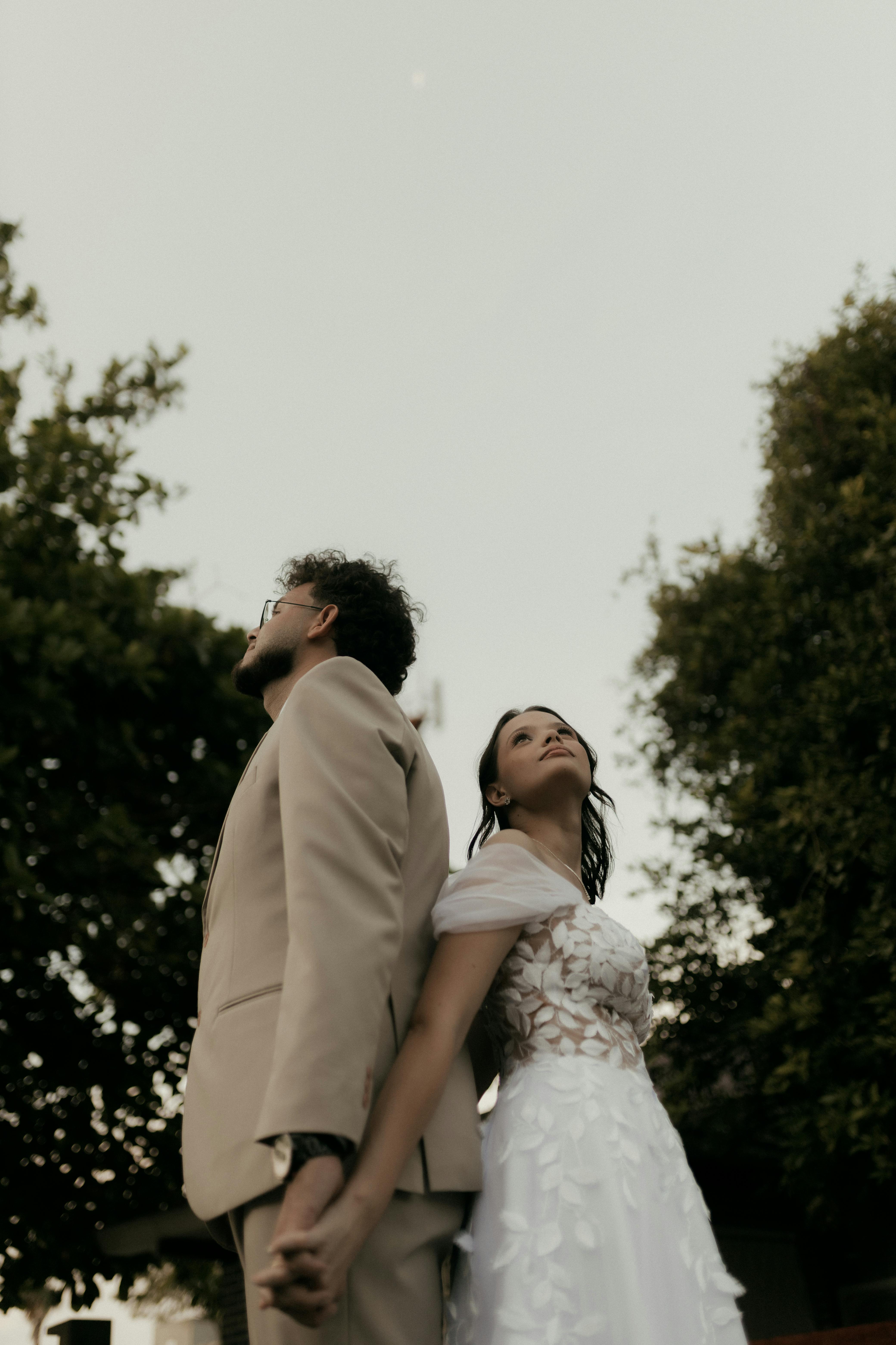 Gratis Una coppia in abiti da sposi si tiene per mano all'aperto al crepuscolo, con lo sguardo rivolto verso l'alto. Foto a disposizione