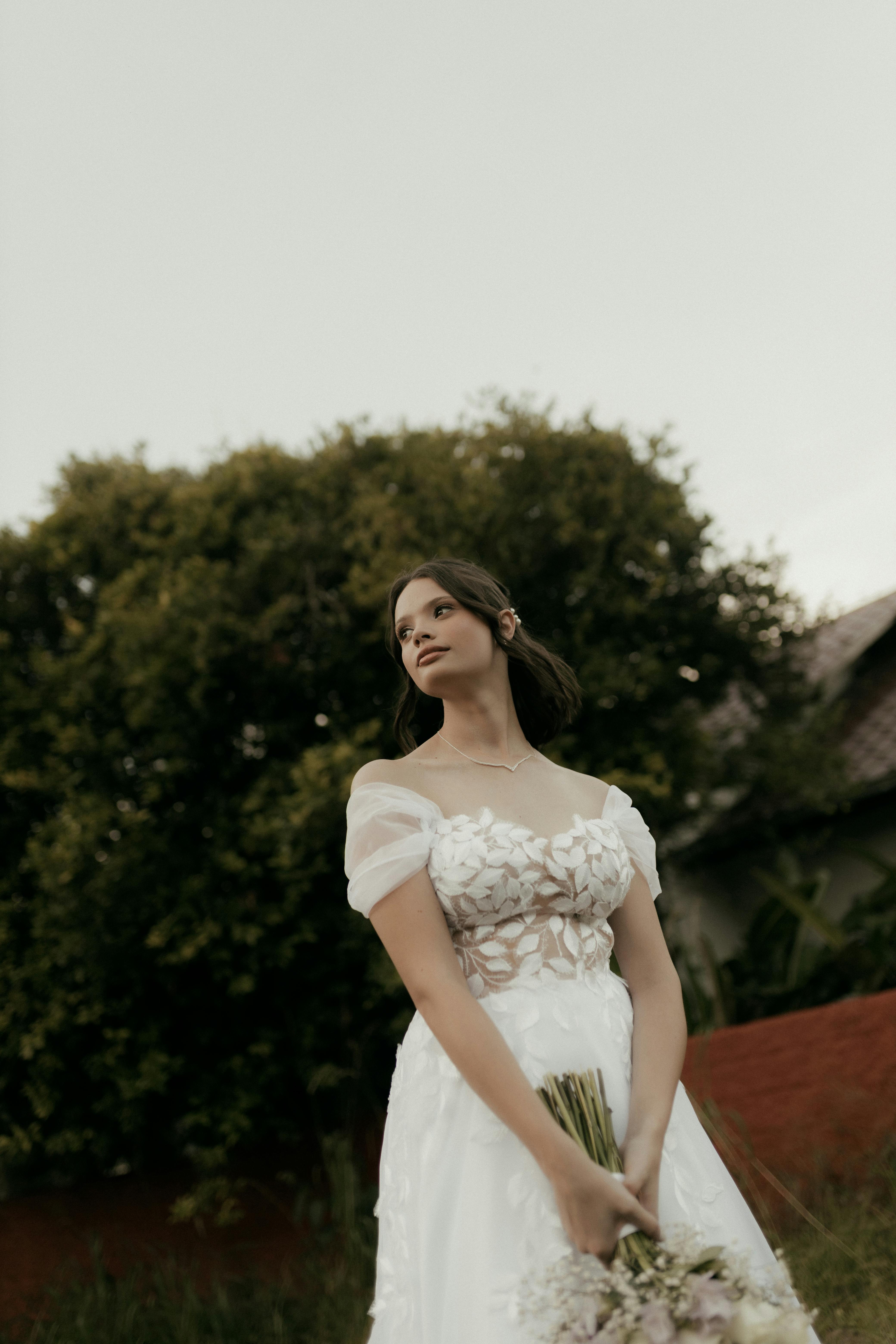 Kostenlos Eine wunderschöne Braut in einem Brautkleid aus Spitze hält einen Brautstrauß im Freien vor dem Hintergrund von Bäumen. Stock-Foto