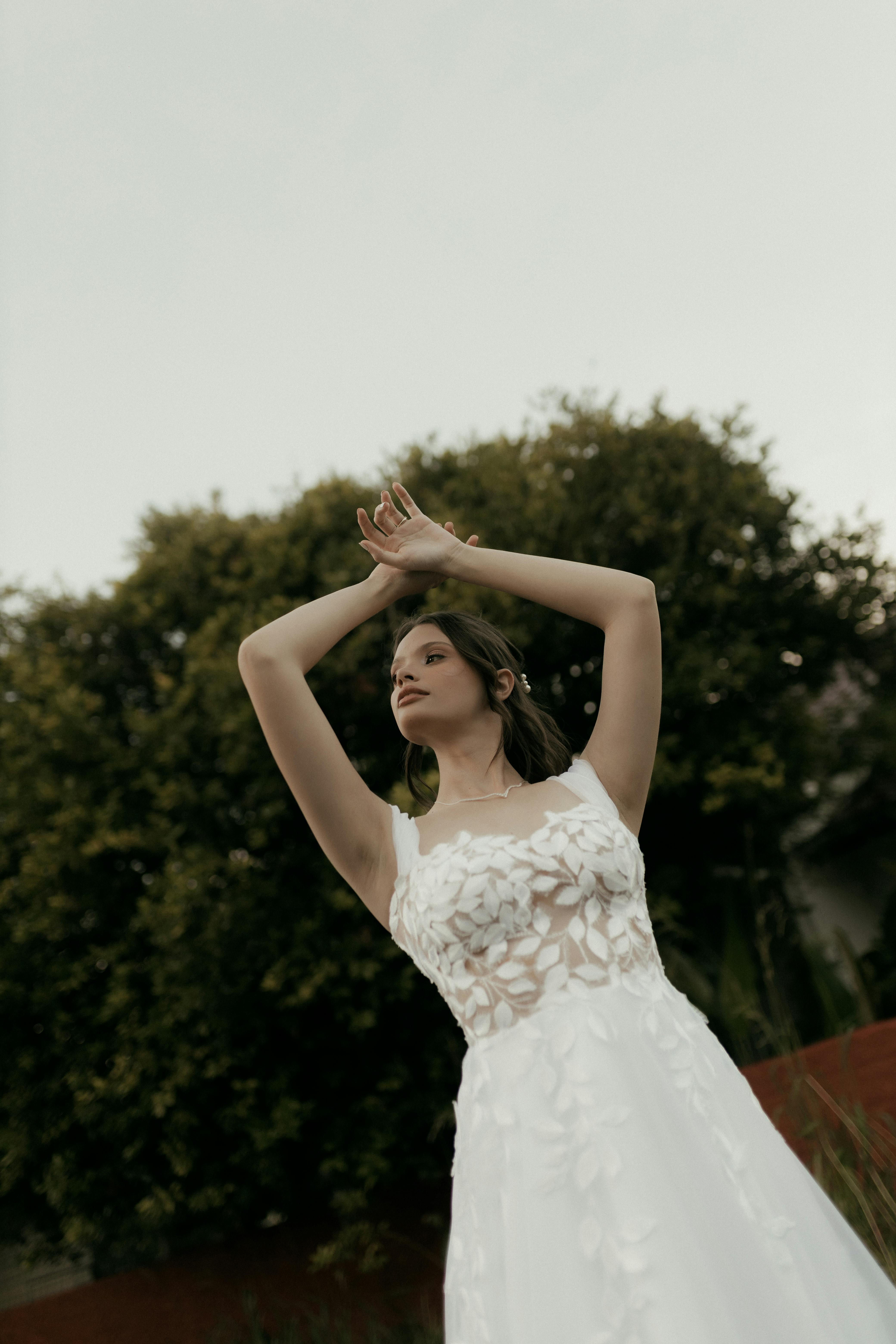 Kostenlos Eine junge Frau posiert anmutig in einem weißen, spitzenbesetzten Brautkleid im Freien, umgeben von Grün. Stock-Foto