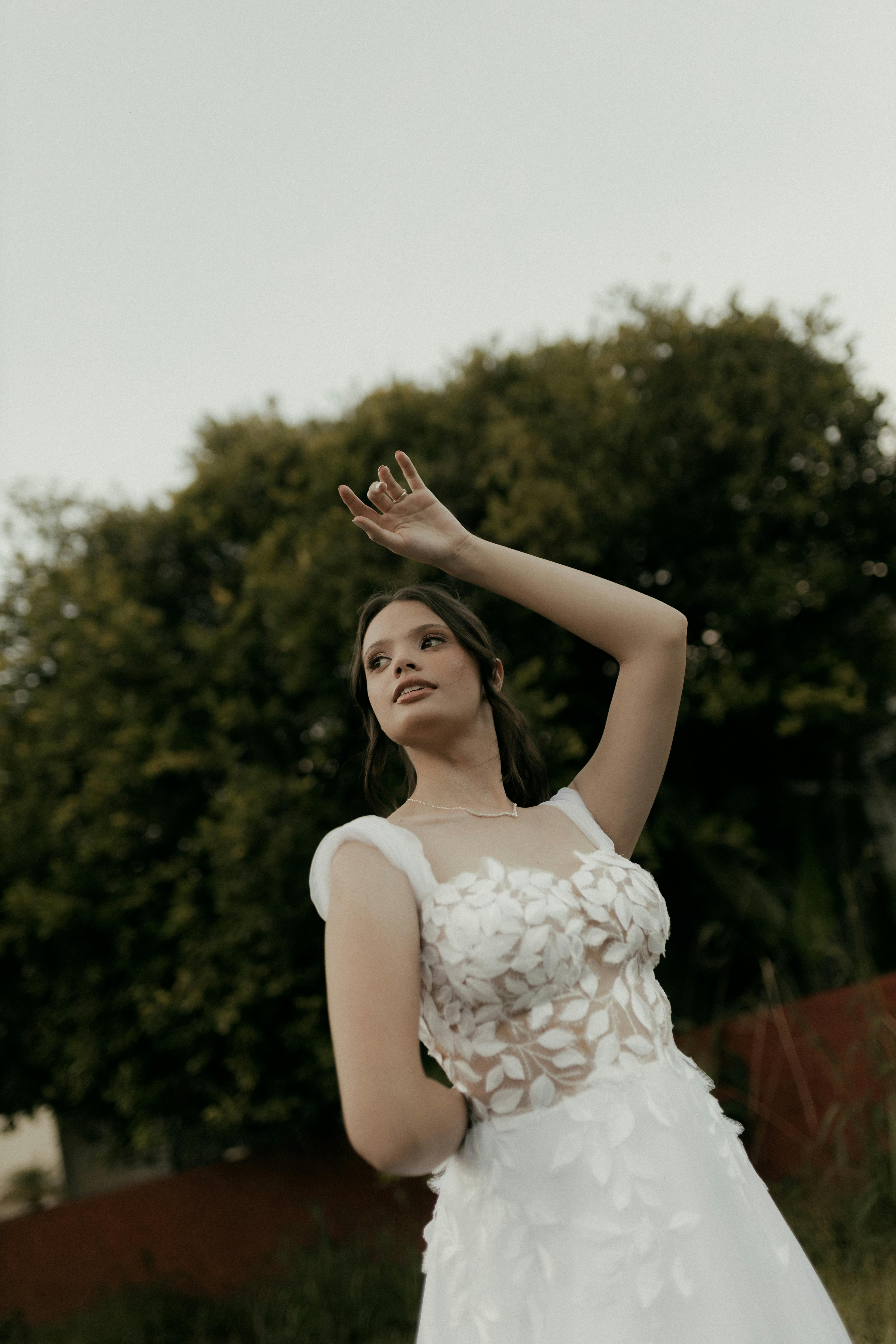 無料 繊細なディテールが施された白いドレスを身にまとった、美しい花嫁が屋外で優雅にポーズをとっている。 写真素材