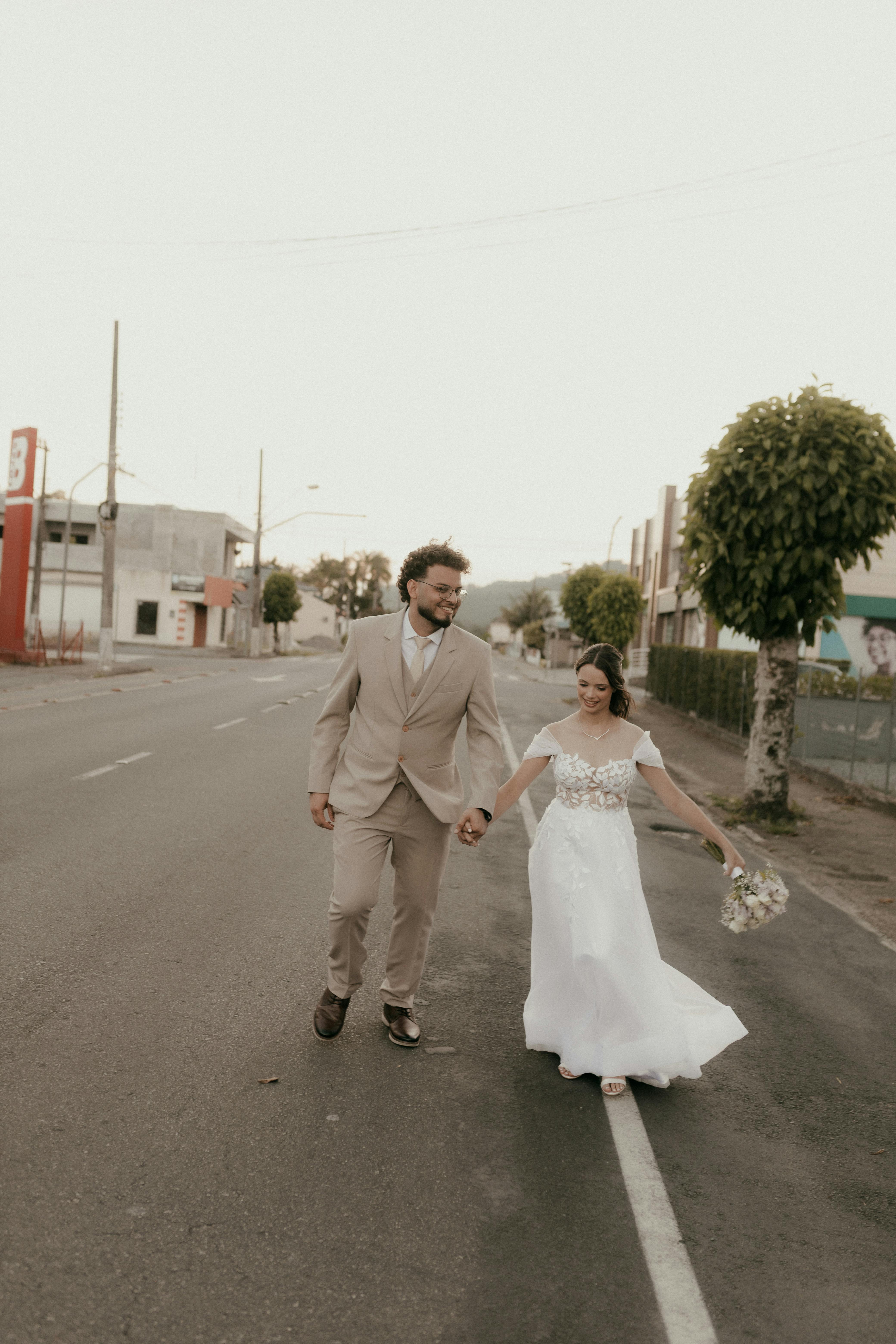 Gratis Gli sposi, in abiti eleganti, camminano mano nella mano per strada in una giornata limpida. Foto a disposizione