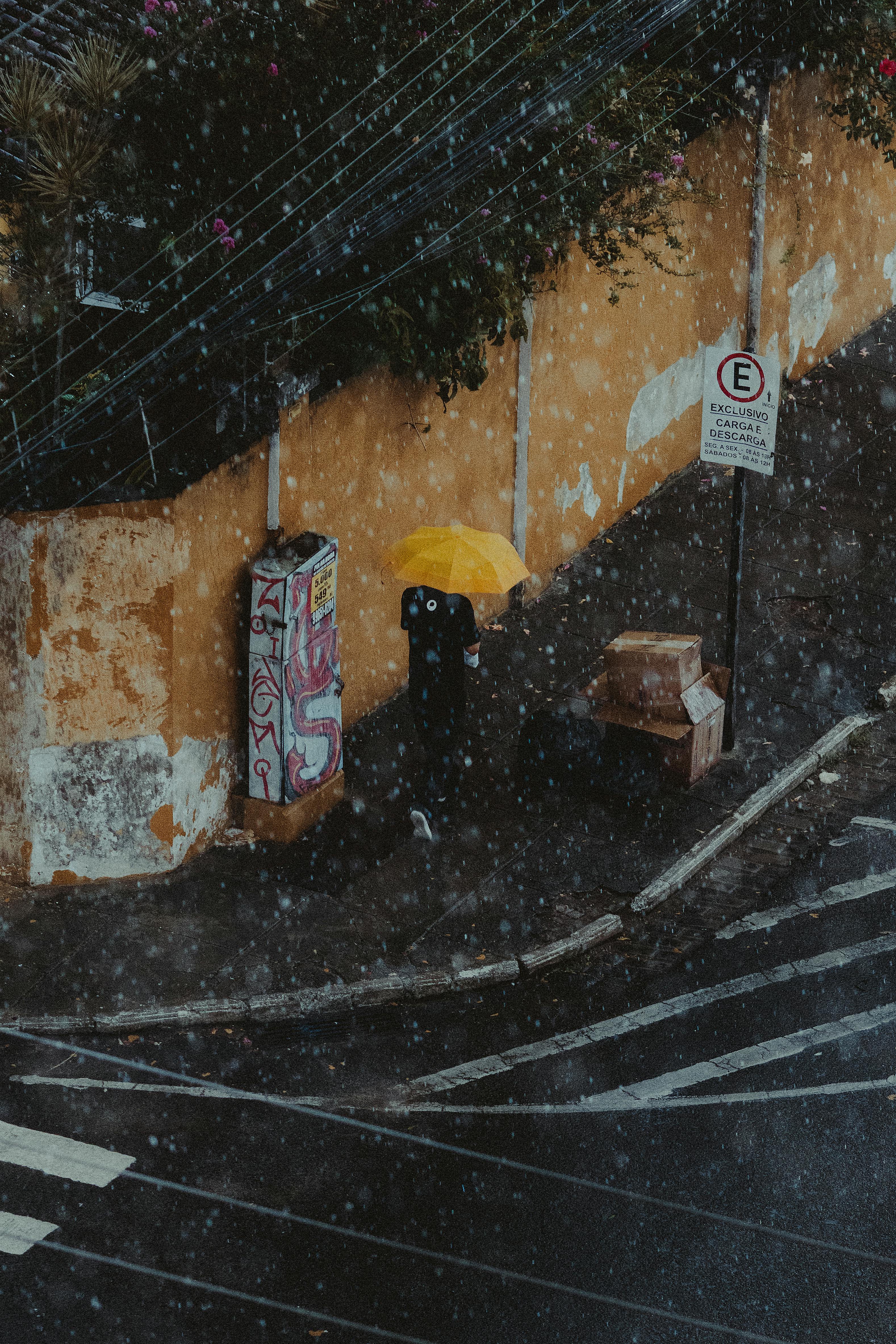 Безкоштовне Дощова вулична сцена з людиною під жовтою парасолькою, що додає яскравого контрасту міському пейзажу. стокове фото