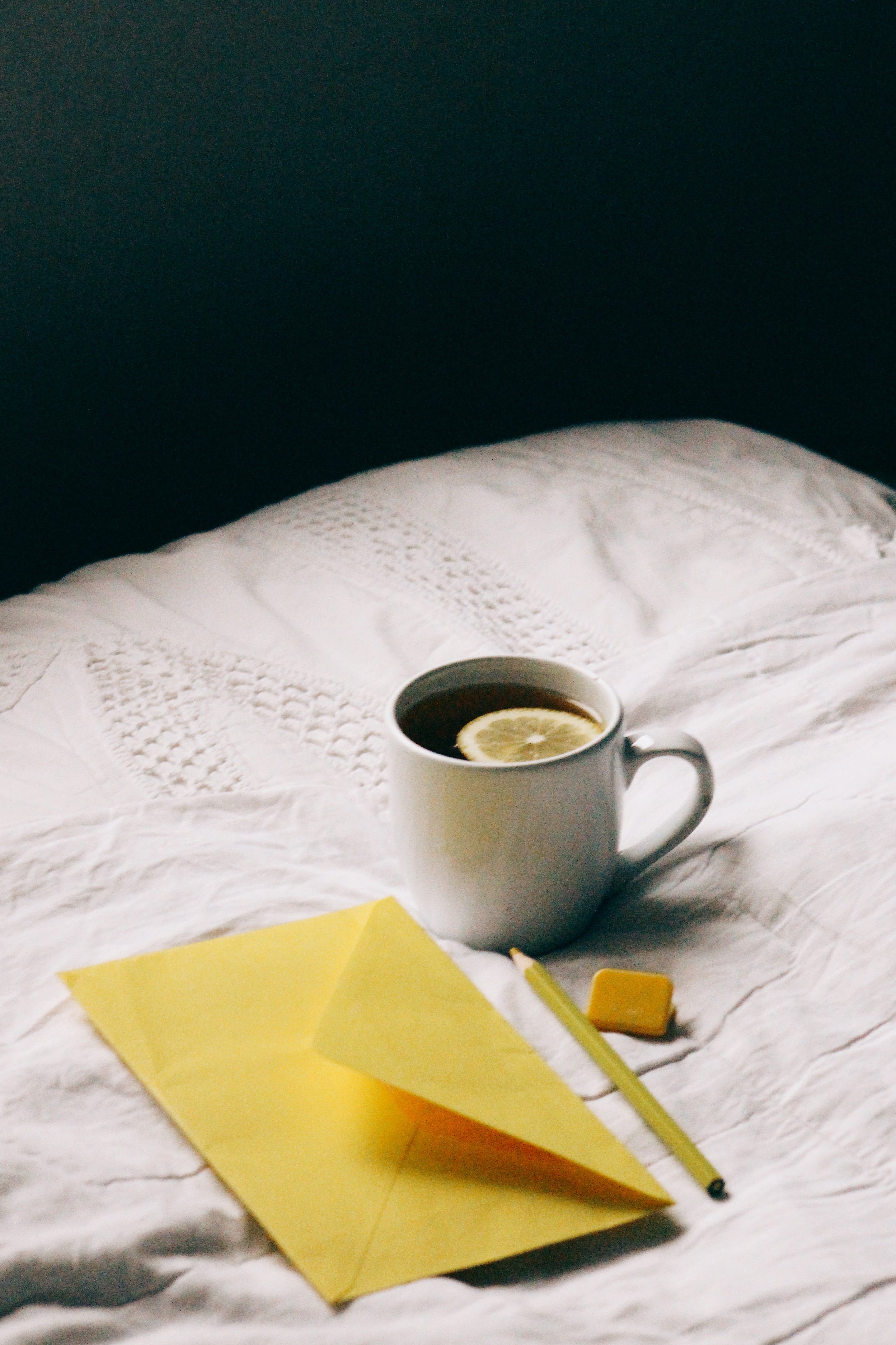 A cozy morning scene with a lemon tea, yellow envelope, and pencil on a white bed.