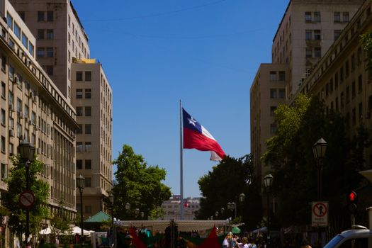 Chilean flag waving between historic buildings in a bustling Santiago city street on a sunny day.