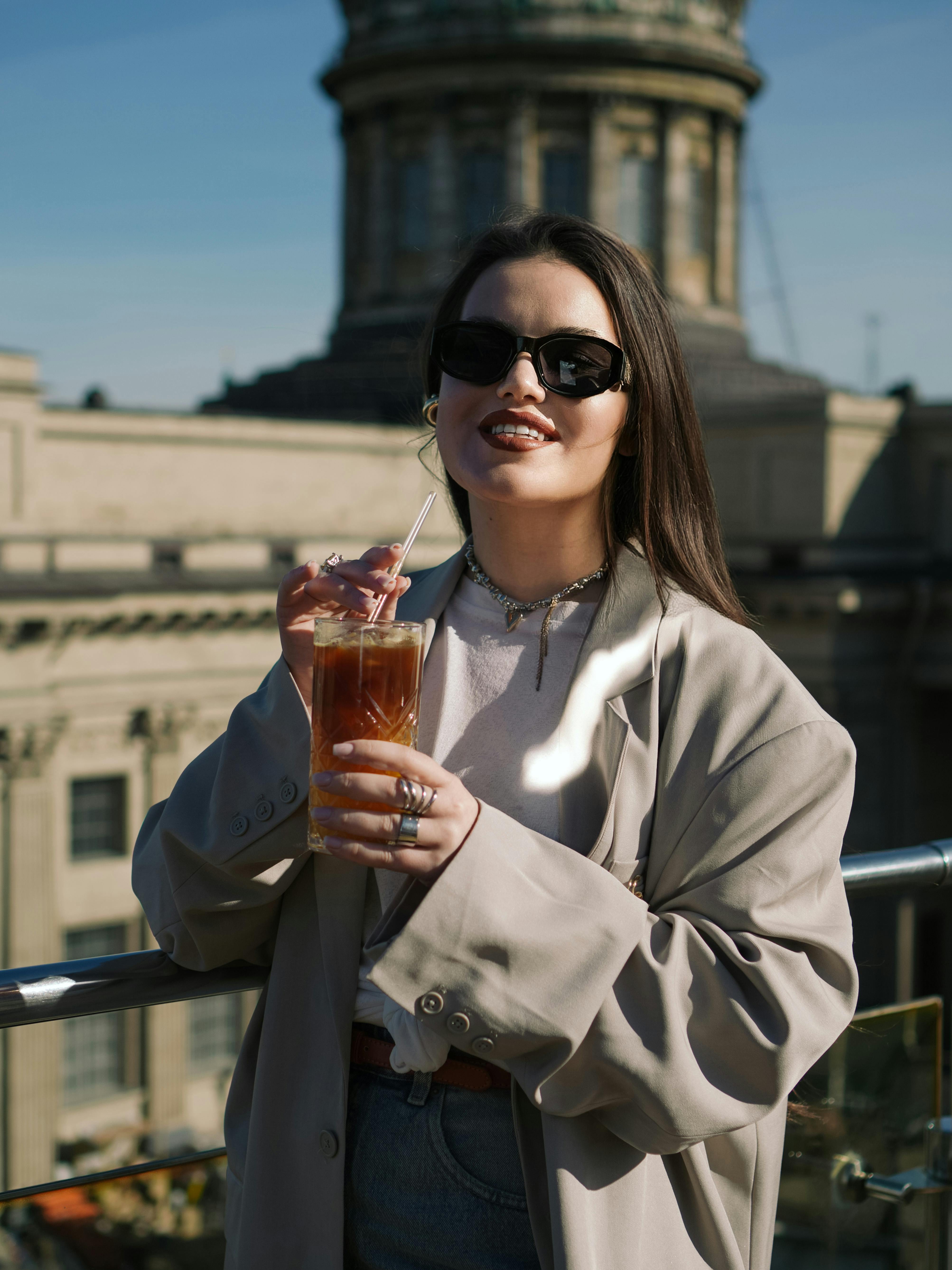 無料 スタイリッシュな女性が、街の建築物を背景に、日当たりの良い屋上で飲み物を楽しんでいる。 写真素材