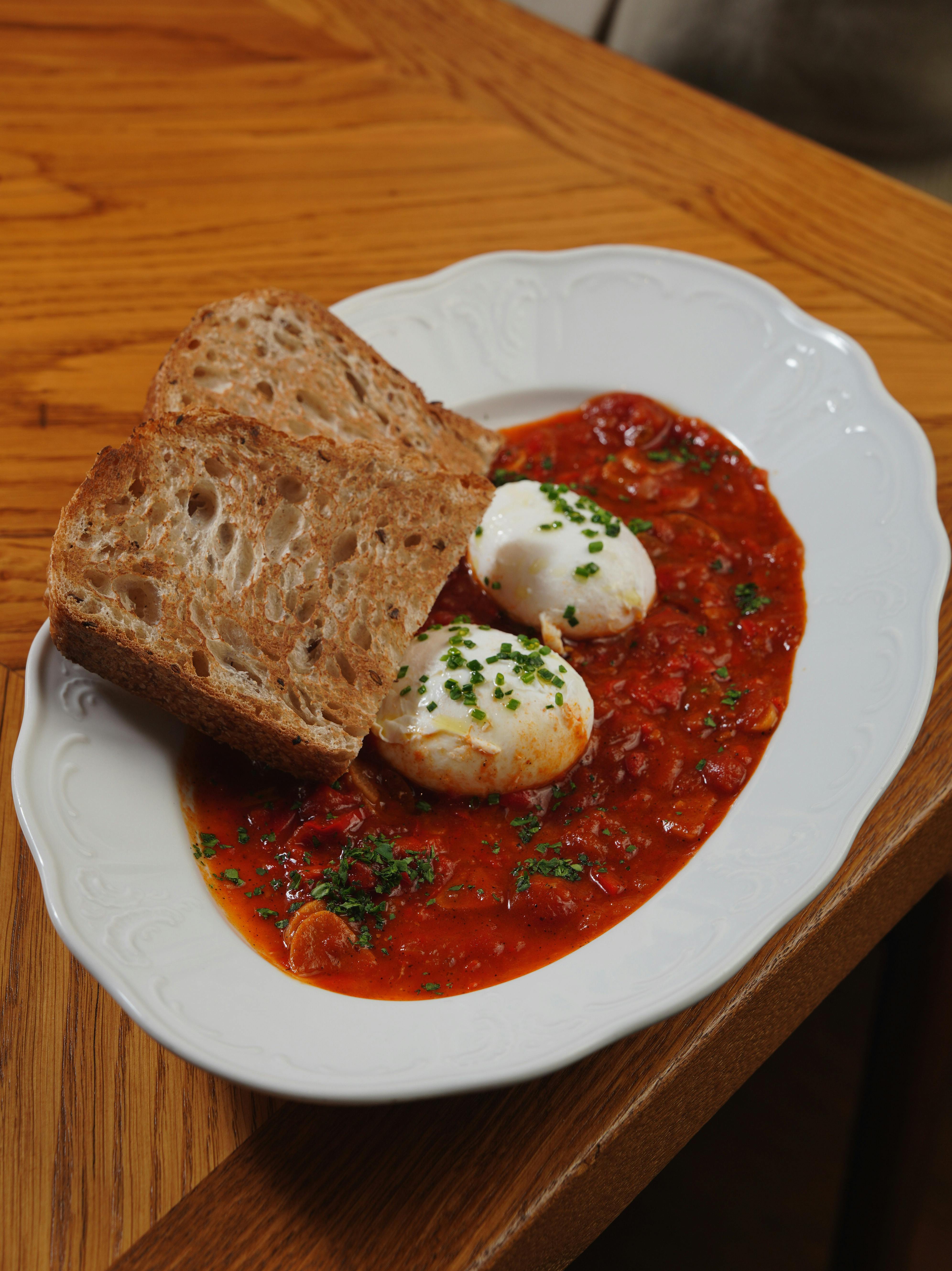 Gratis Un piatto di shakshuka con uova in camicia e pane rustico su un tavolo di legno. Foto a disposizione