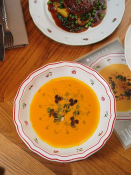 Gourmet pumpkin soup garnished with seeds and herbs in elegant floral bowl on wooden table.