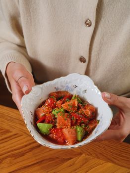 Close-up of a fresh salmon and avocado poke bowl held by a person in beige attire.