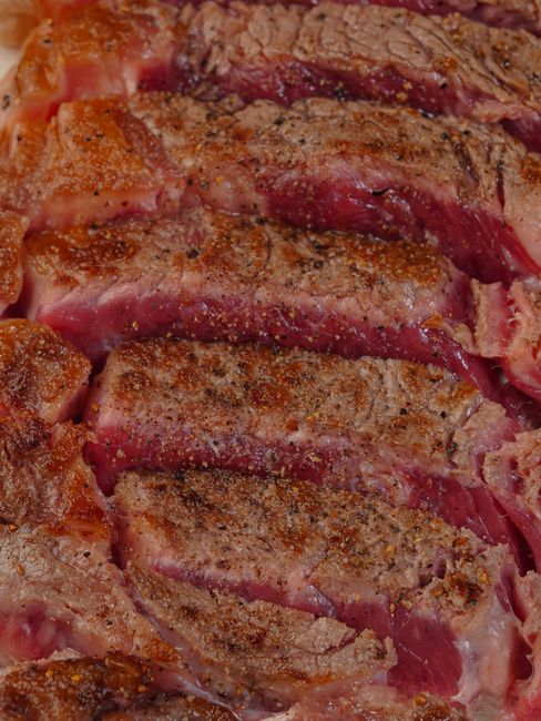 A thick ribeye steak sliced open on a wooden board, showing an even pink center edge to edge and a dark brown crust, with a cast iron skillet and rosemary sprig in the background.