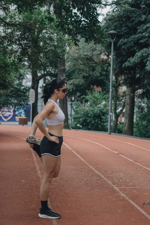 Free Fit woman stretches leg on track surrounded by trees, preparing for a run outdoors. Stock Photo