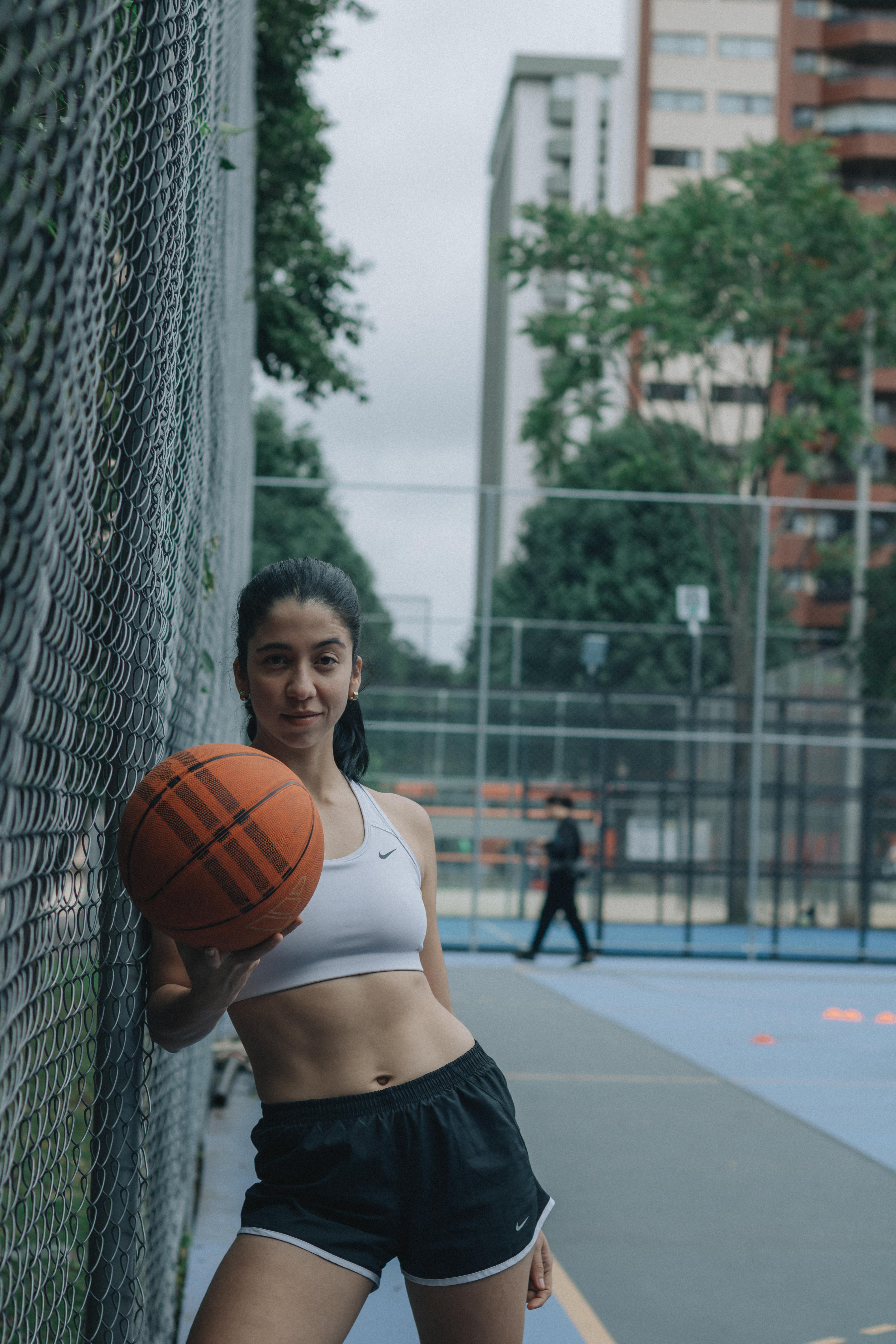 免費 一位身材健美的女子在戶外城市球場上擺出籃球姿勢，展現了她的健康體魄和時尚品味。 圖庫相片