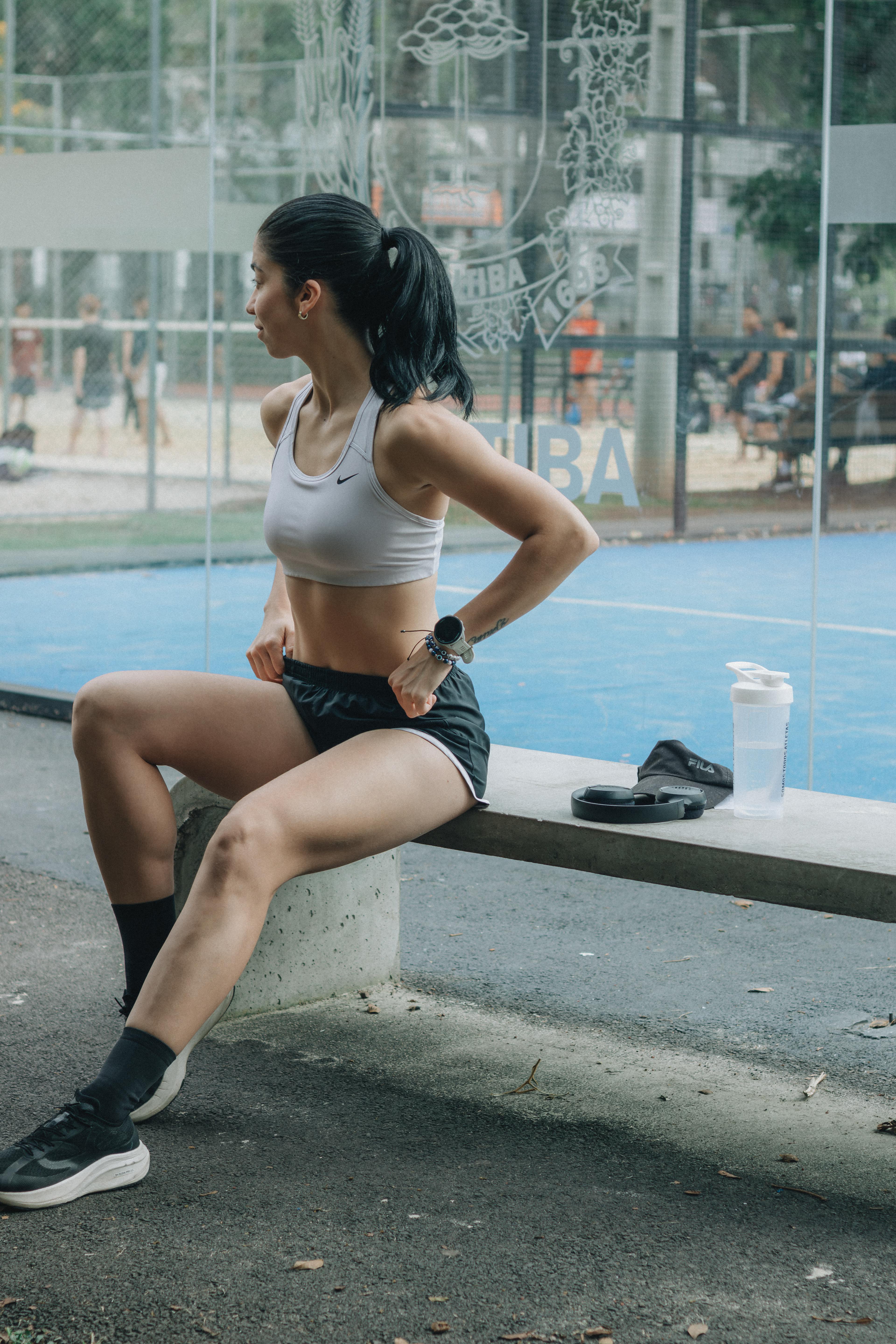 ฟรี นักกีฬาหญิงกำลังพักผ่อนบนม้านั่งกลางแจ้ง ในสภาพแวดล้อมกีฬากลางวันที่มีแสงสว่างสดใส คลังภาพถ่าย