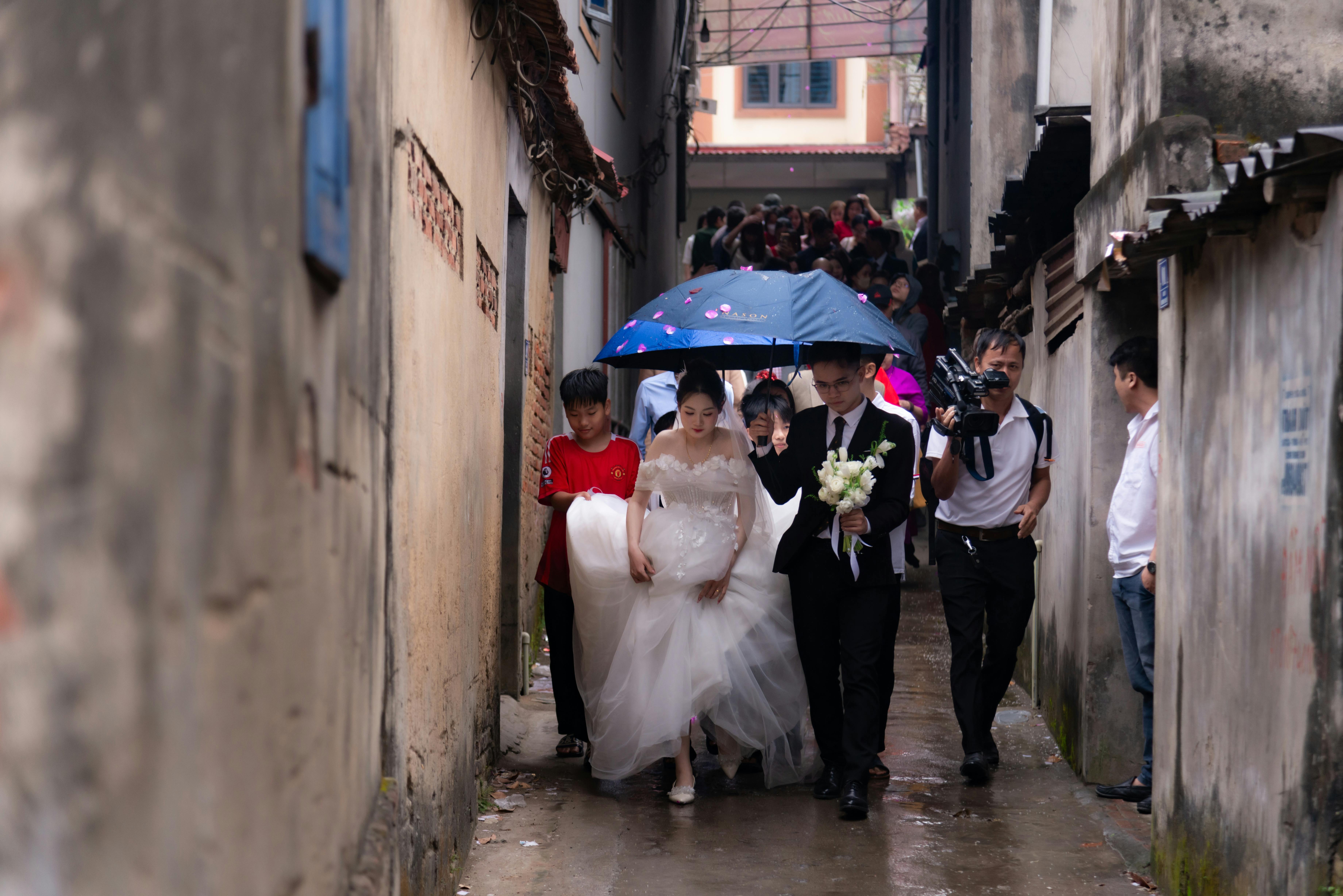 Gratis Los novios caminan bajo un paraguas por un callejón estrecho con su séquito nupcial en un día lluvioso. Foto de stock