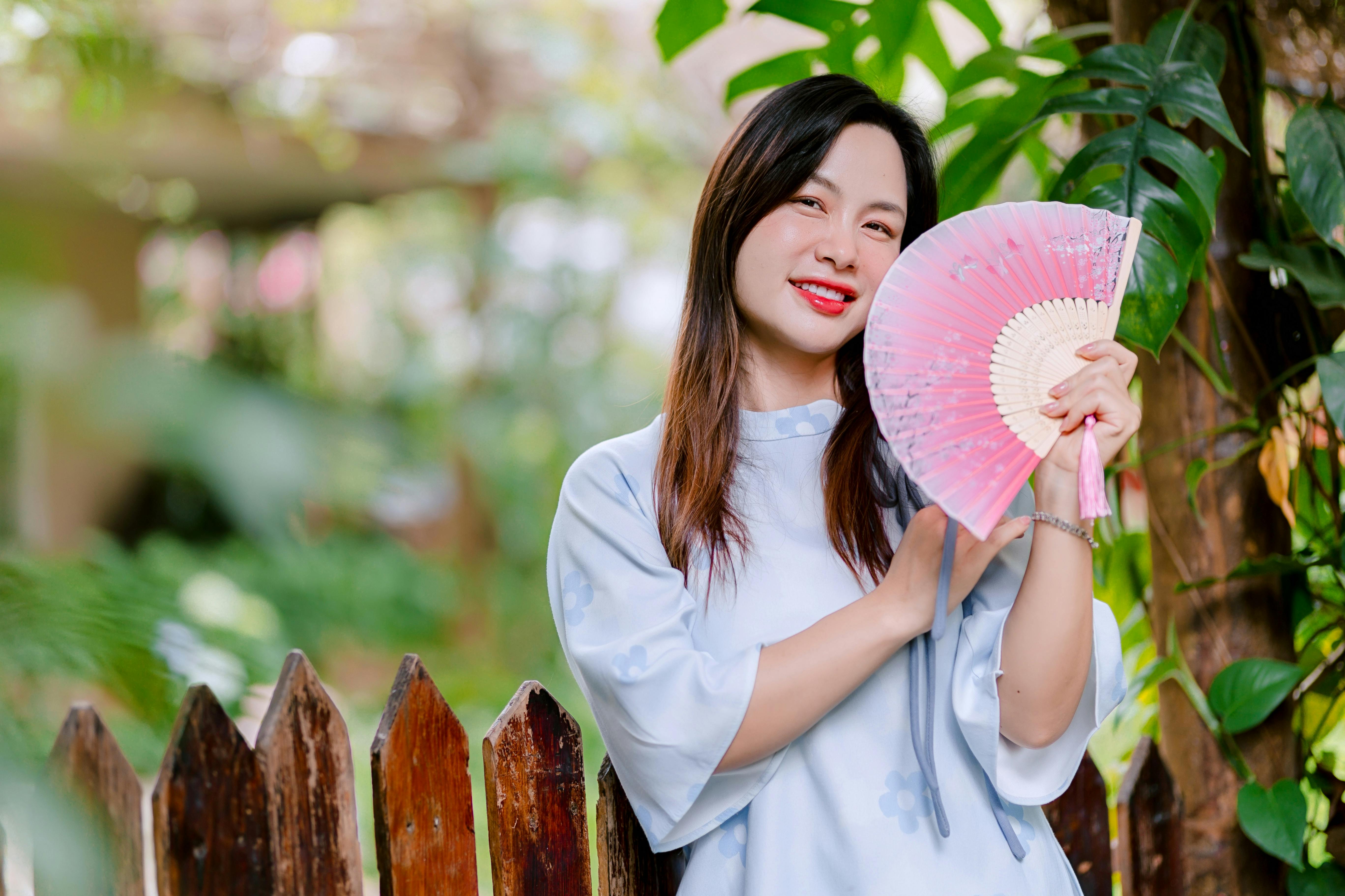 Ücretsiz Yeşilliklerle dolu bir bahçede, açık havada duran, mavi elbiseli neşeli bir kadın elinde pembe bir yelpaze tutuyor. Stok Fotoğraflar