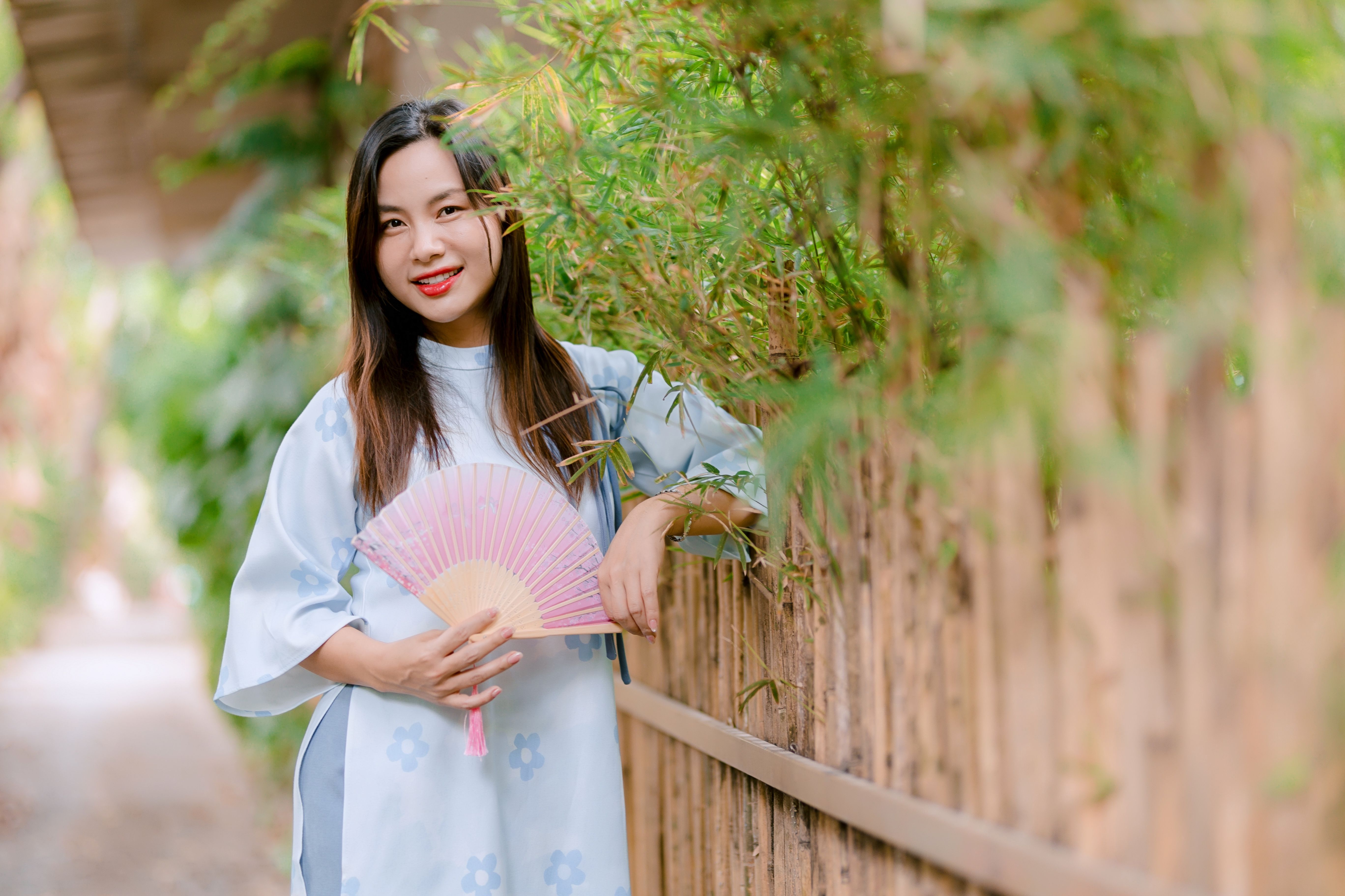 Woman in traditional attire holds bamboo fan in a serene outdoor setting.
