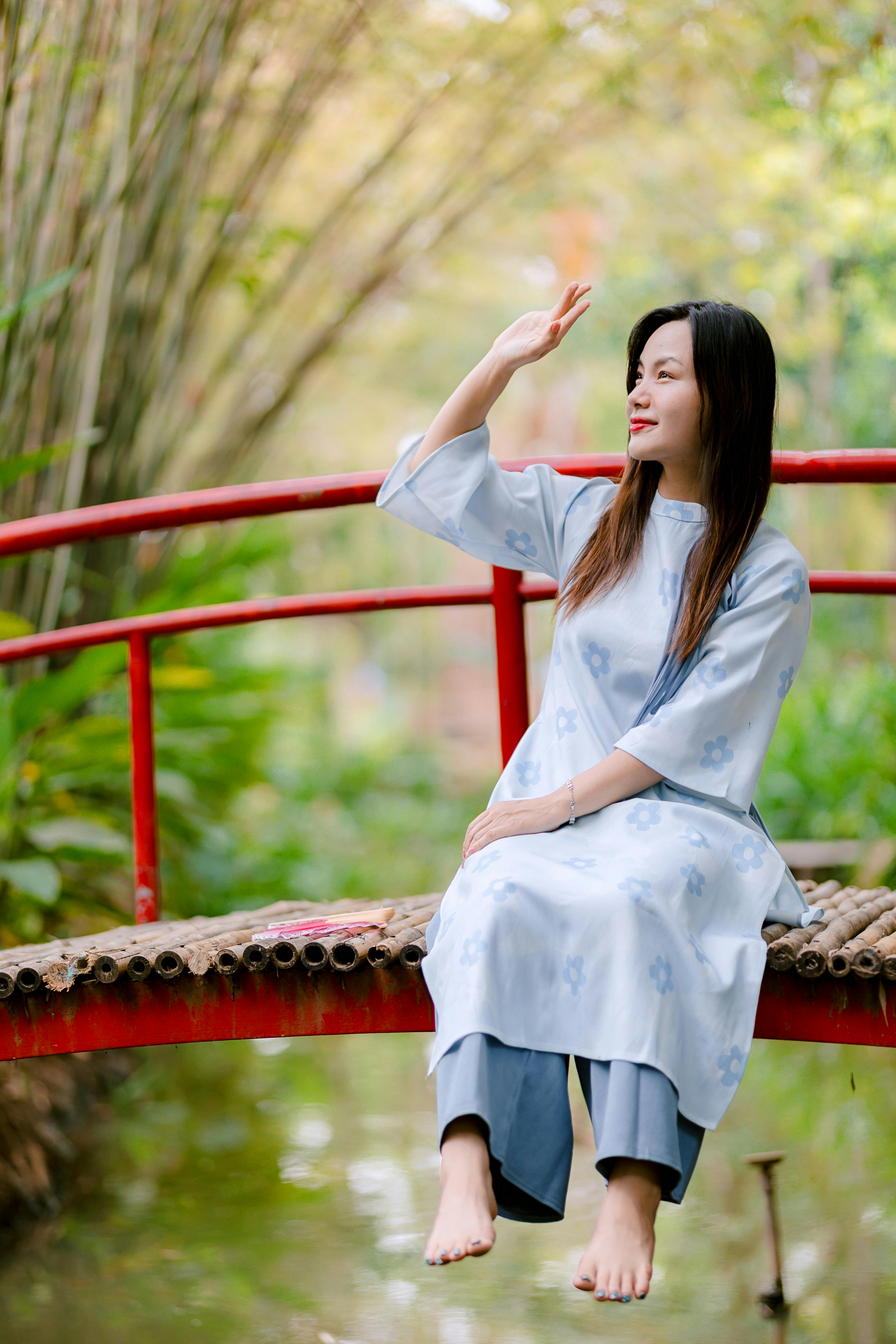 Ücretsiz Geleneksel kıyafetler giymiş bir kadın, yemyeşil bitki örtüsüyle çevrili bambu bir köprü üzerinde oturuyor. Stok Fotoğraflar