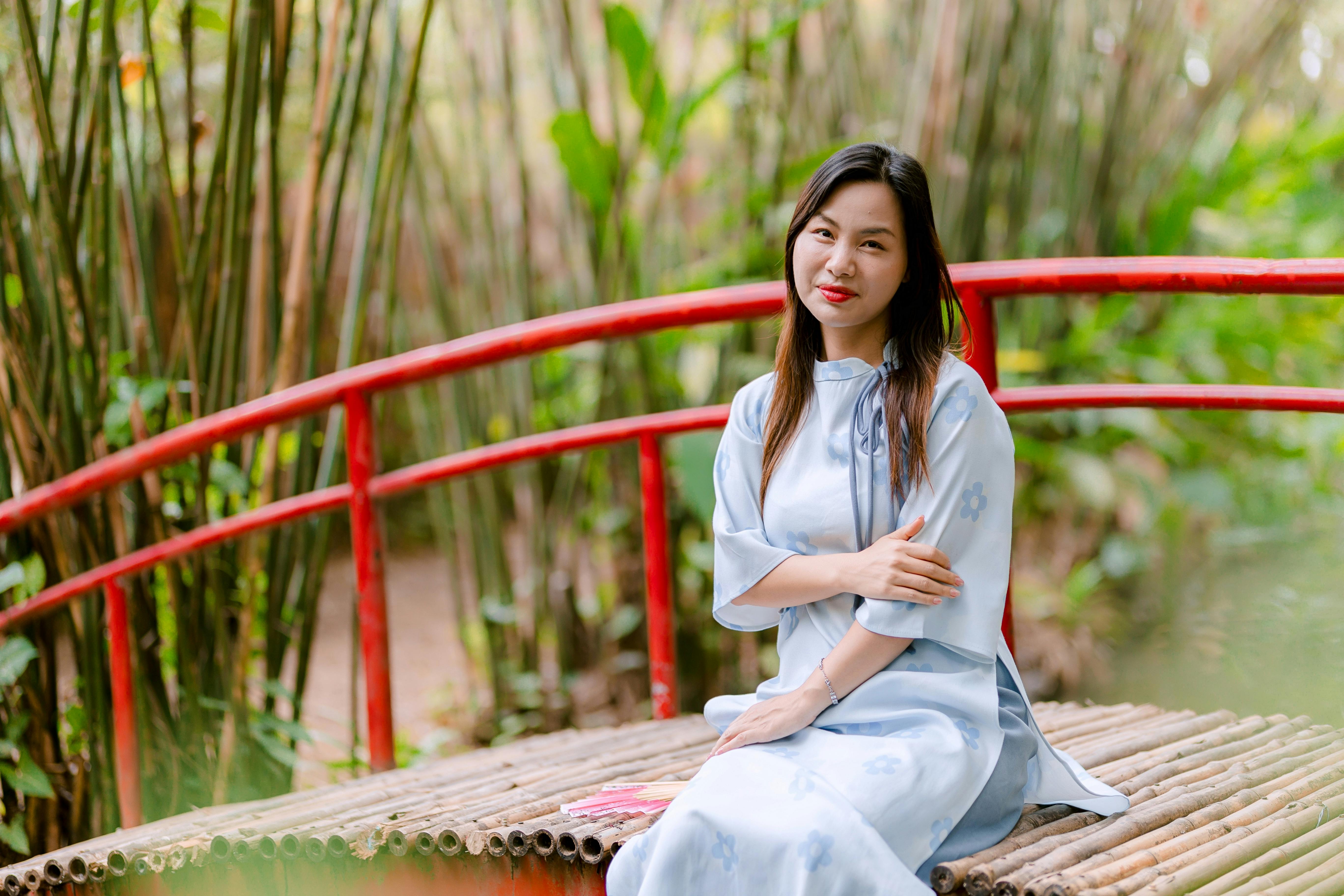 Gratis Una donna in abito tradizionale siede serenamente su un ponte rosso, circondata da bambù e piante rigogliose. Foto a disposizione