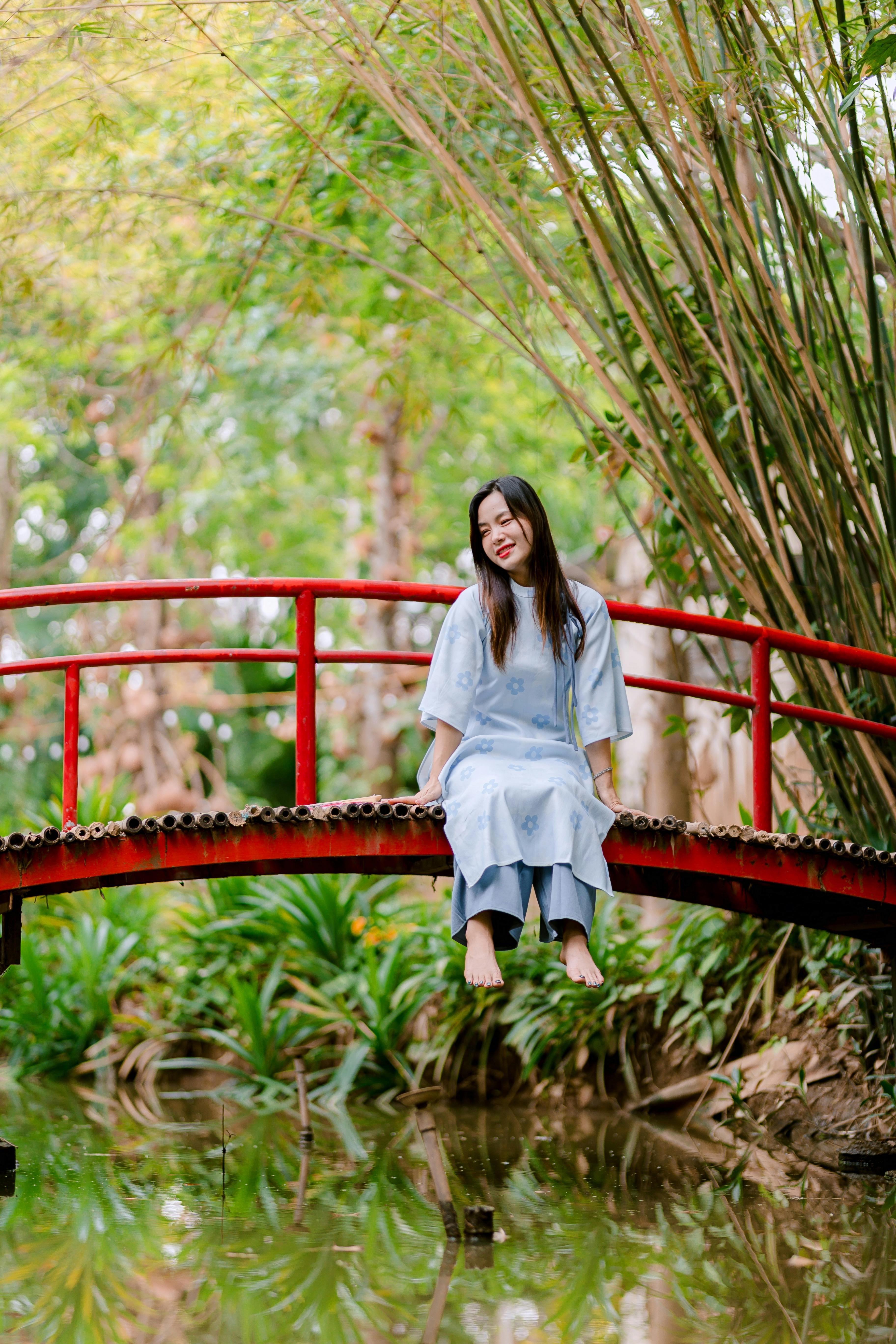 Gratis Kvinde sidder fredeligt på en rød havebro over en dam midt i frodigt grønt. Lagerfoto
