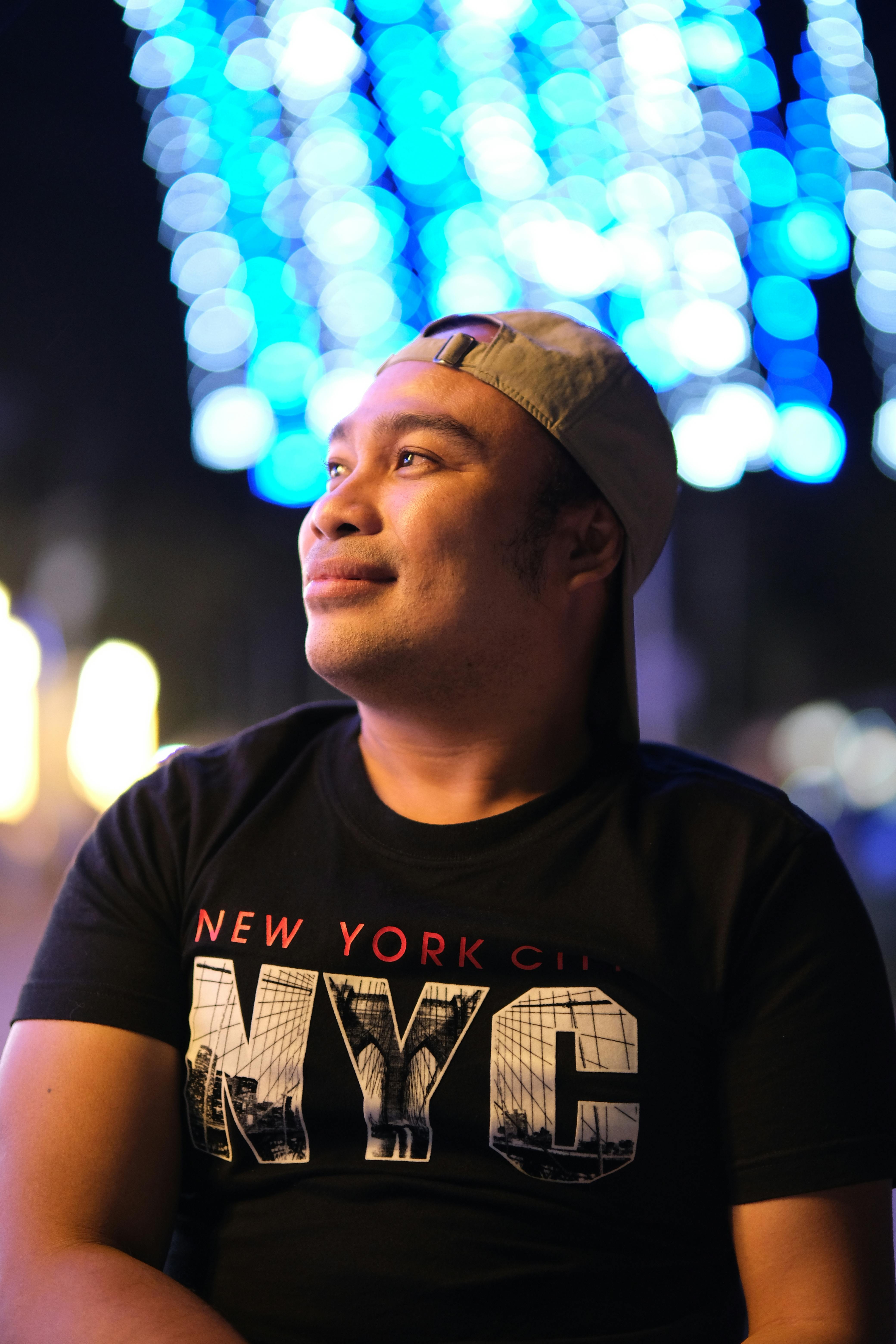 A man in a cap enjoys the vibrant city lights at night wearing a NYC t-shirt.