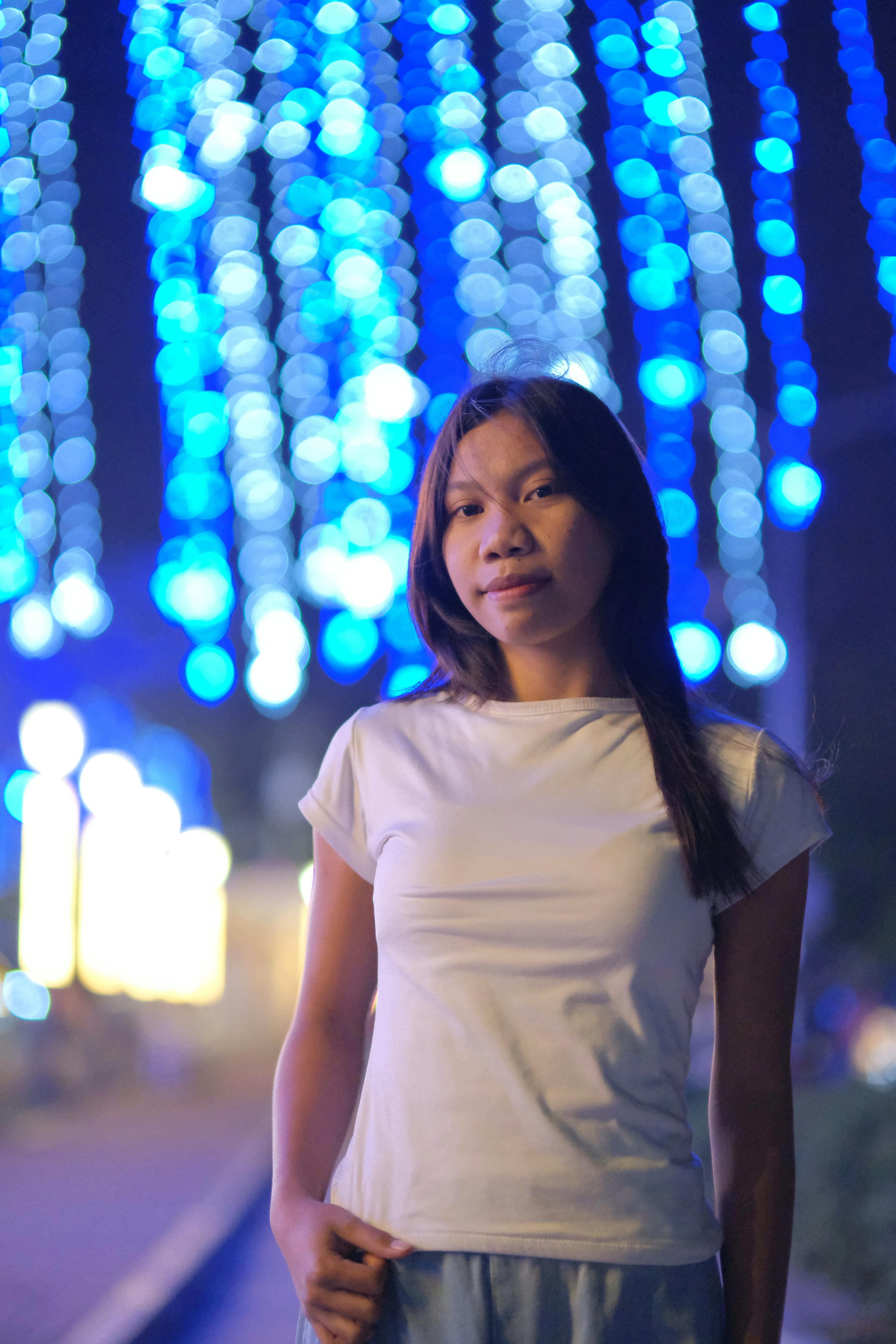 Kostenlos Porträt einer jungen Frau, die nachts unter hellblauen Lichterketten steht. Stock-Foto