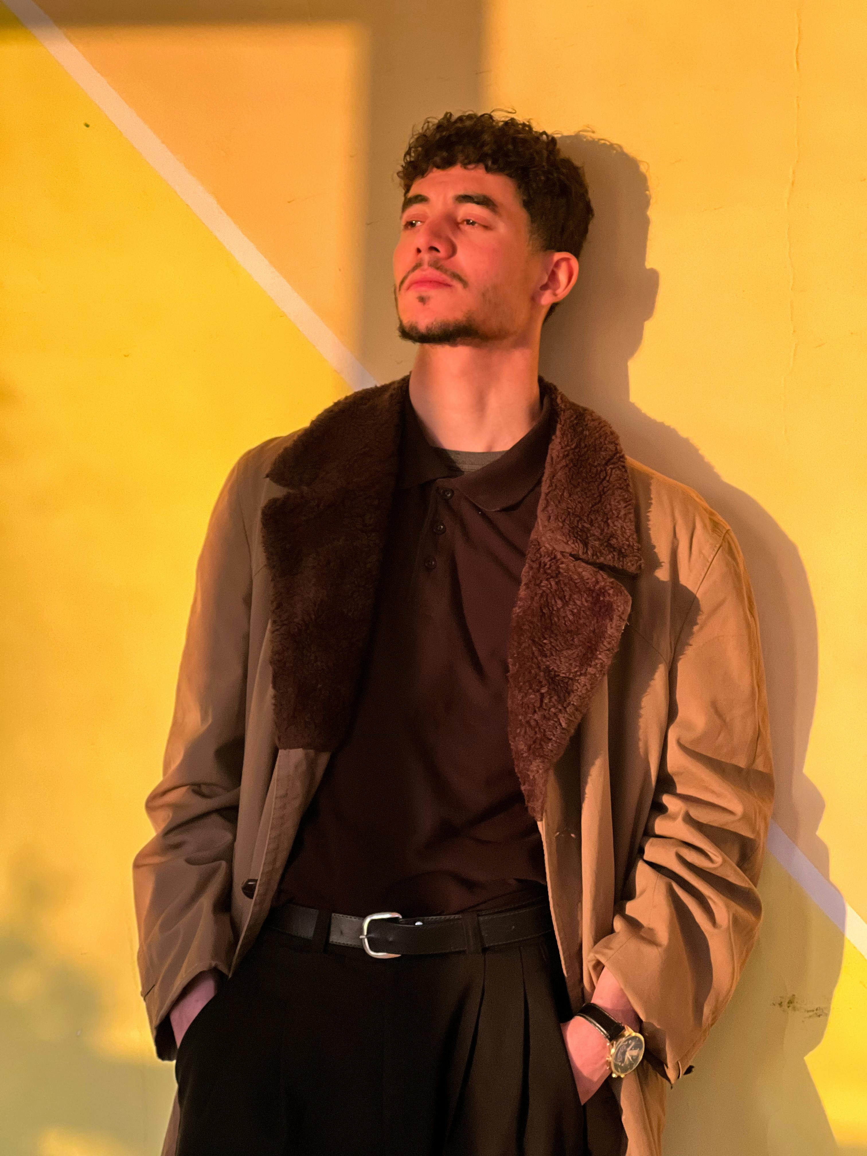 Gratuit Un jeune homme en manteau marron se prélasse au soleil devant un mur jaune chaud, incarnant à merveille le style automnal. Photos