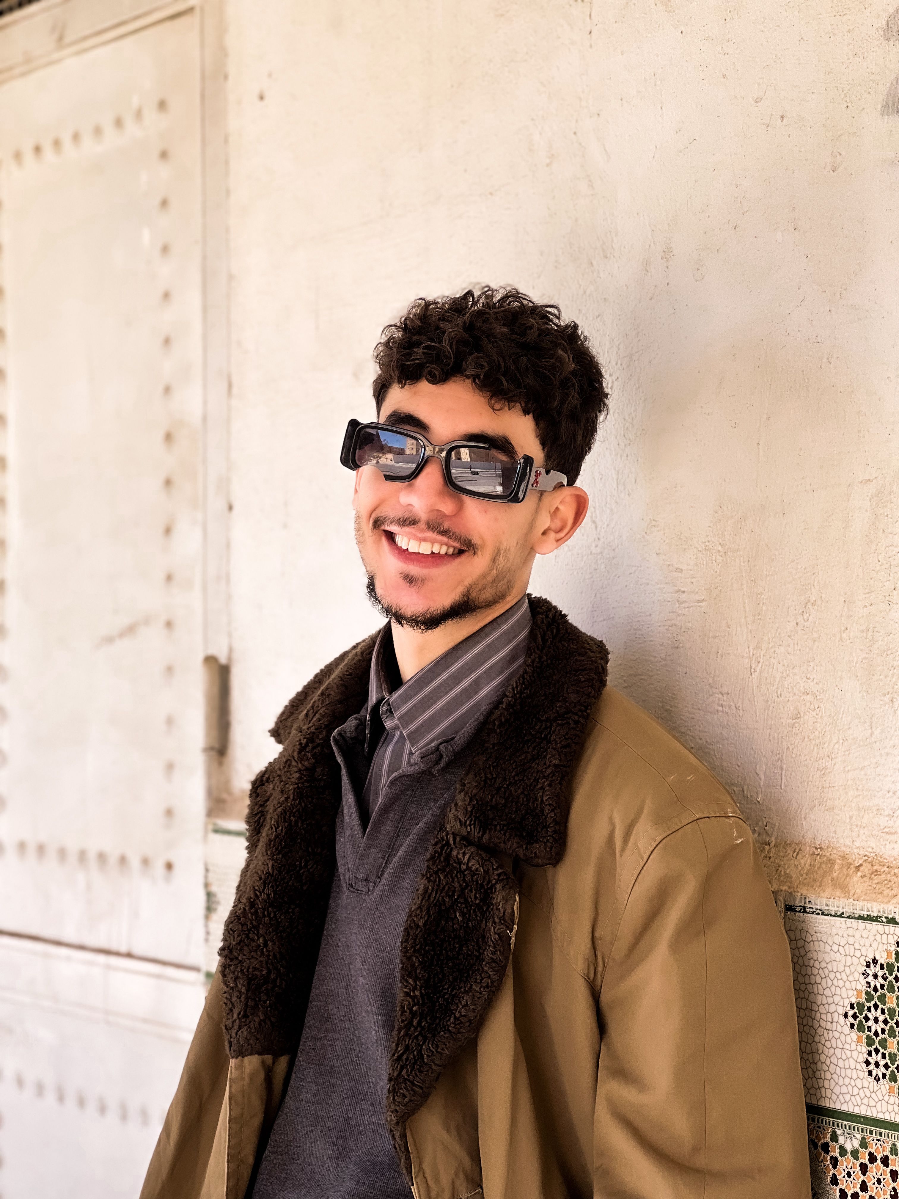 Young man smiling casually in Laghouat, Algeria, wearing sunglasses and a coat.