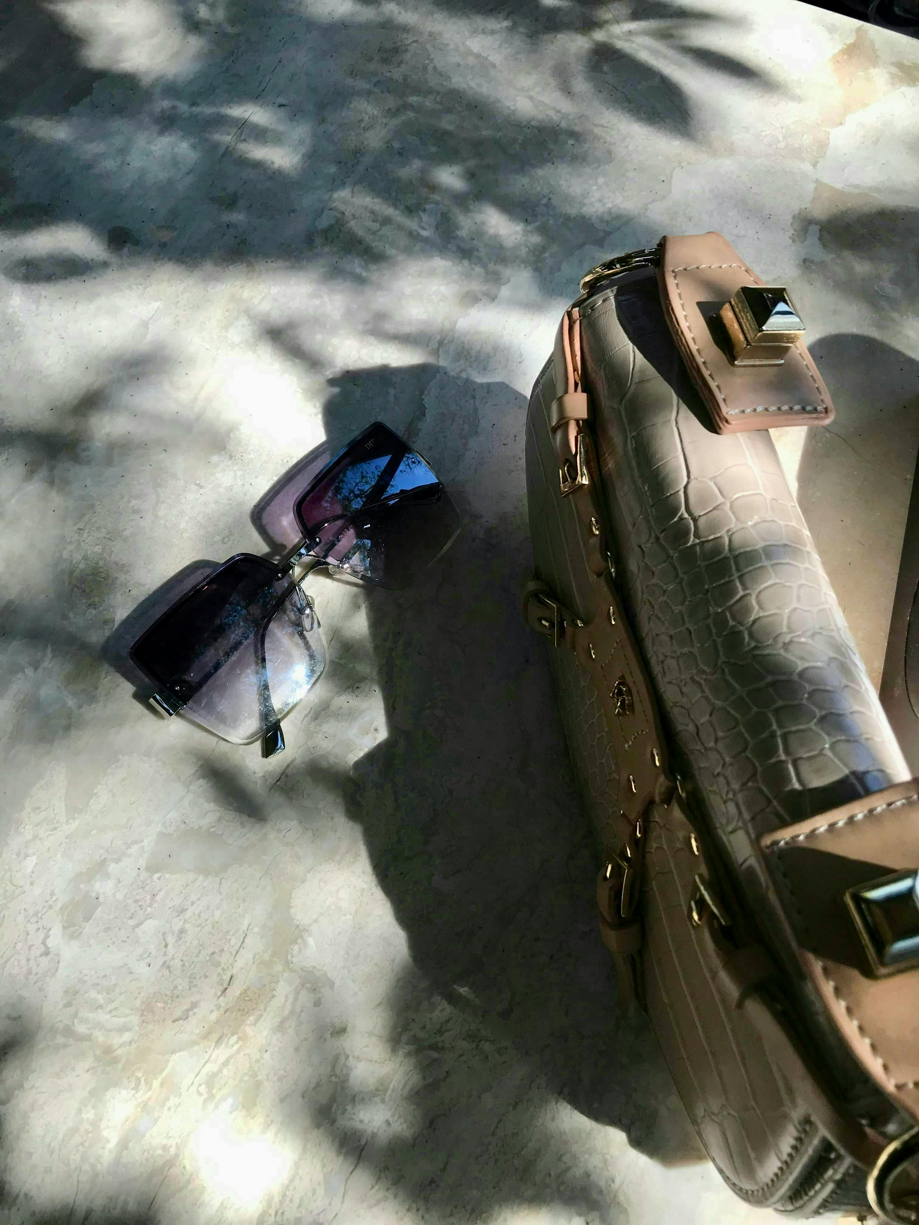 Gratis Occhiali da sole di alta moda e borsa di lusso su una superficie ruvida alla luce del sole. Foto a disposizione