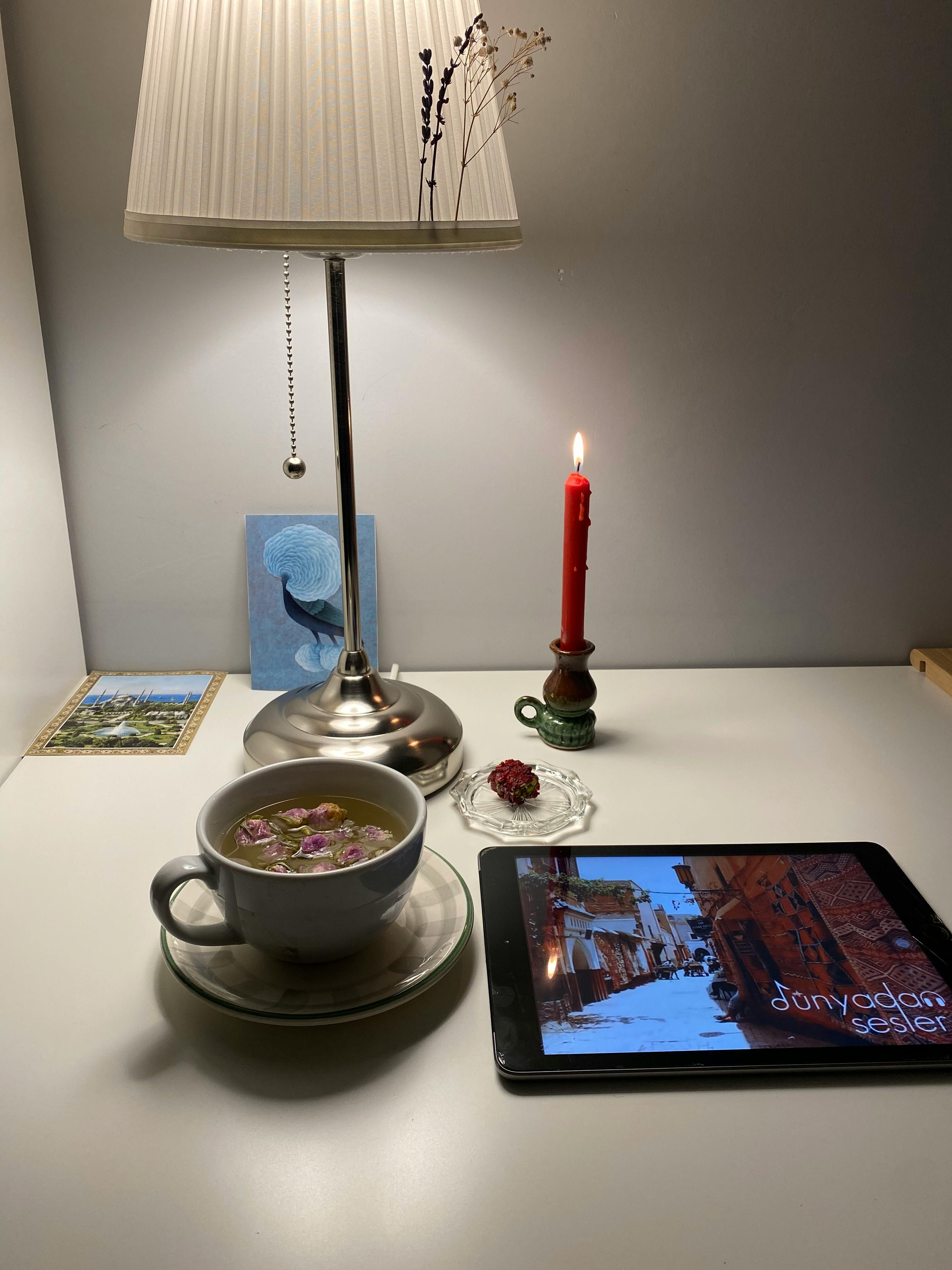 Gratis Un ambiente interno accogliente con una tazza da tè, un tablet, una lampada e una candela su una scrivania. Foto a disposizione
