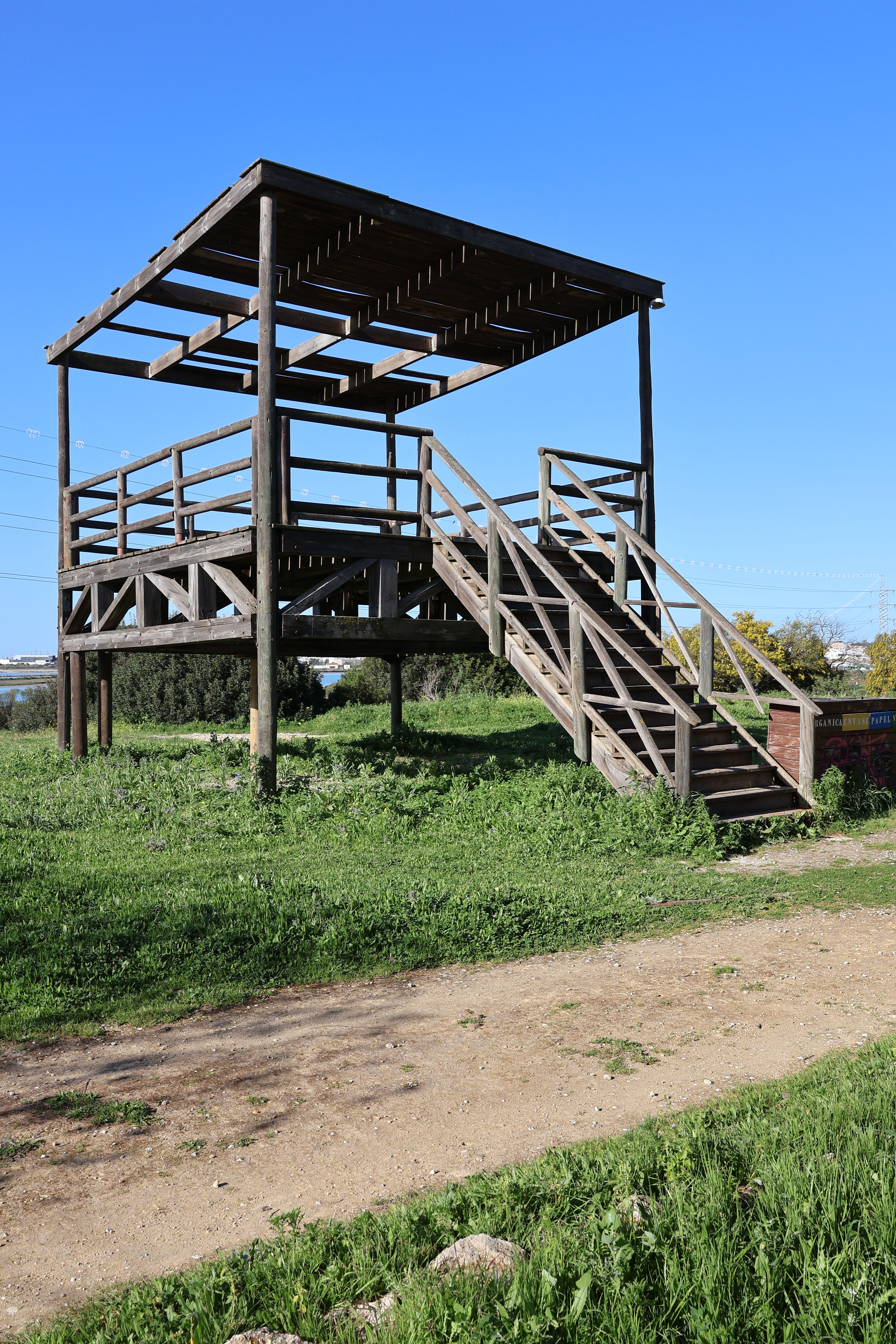 A wooden observation deck in a grassy area under a clear blue sky in El Puerto de Santa María, Spain.