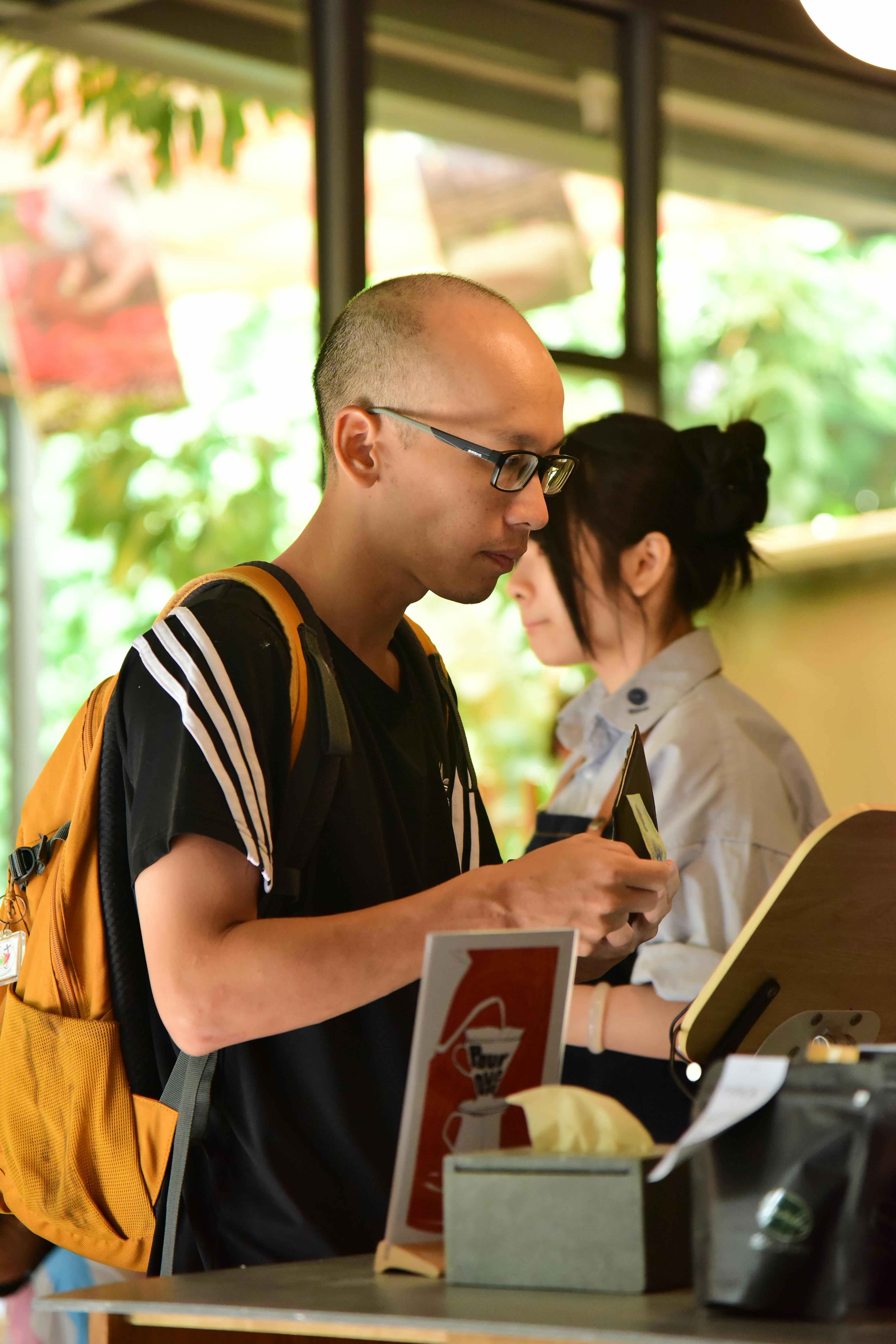 De franc Un home vestit de manera informal amb una motxilla fa una comanda al taulell d'una cafeteria; l'escena captura la vida quotidiana. Foto d'estoc
