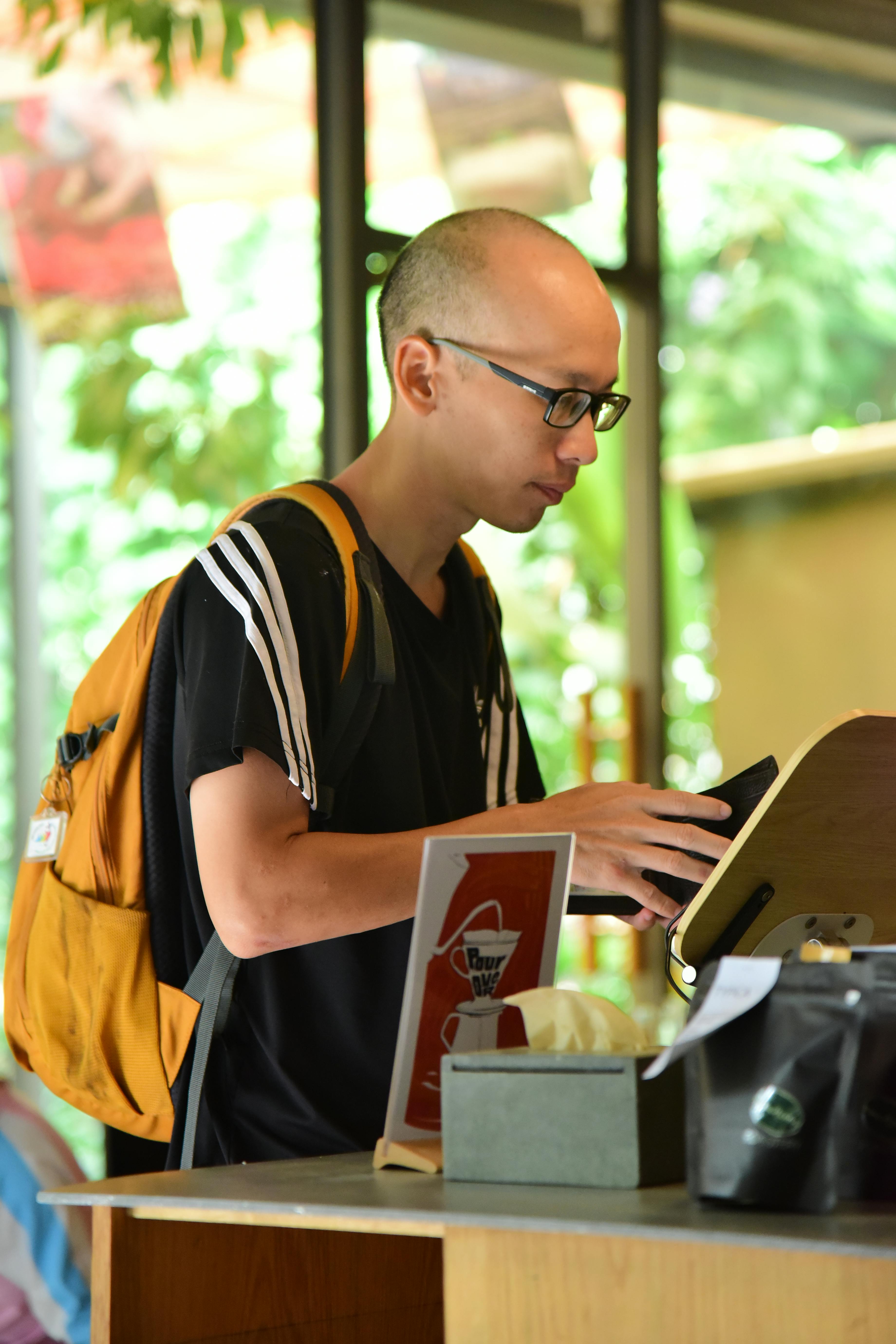 無料 リュックサックを背負った若い男性が、外に緑が見えるカフェのカウンターでデバイスを使用している。 写真素材