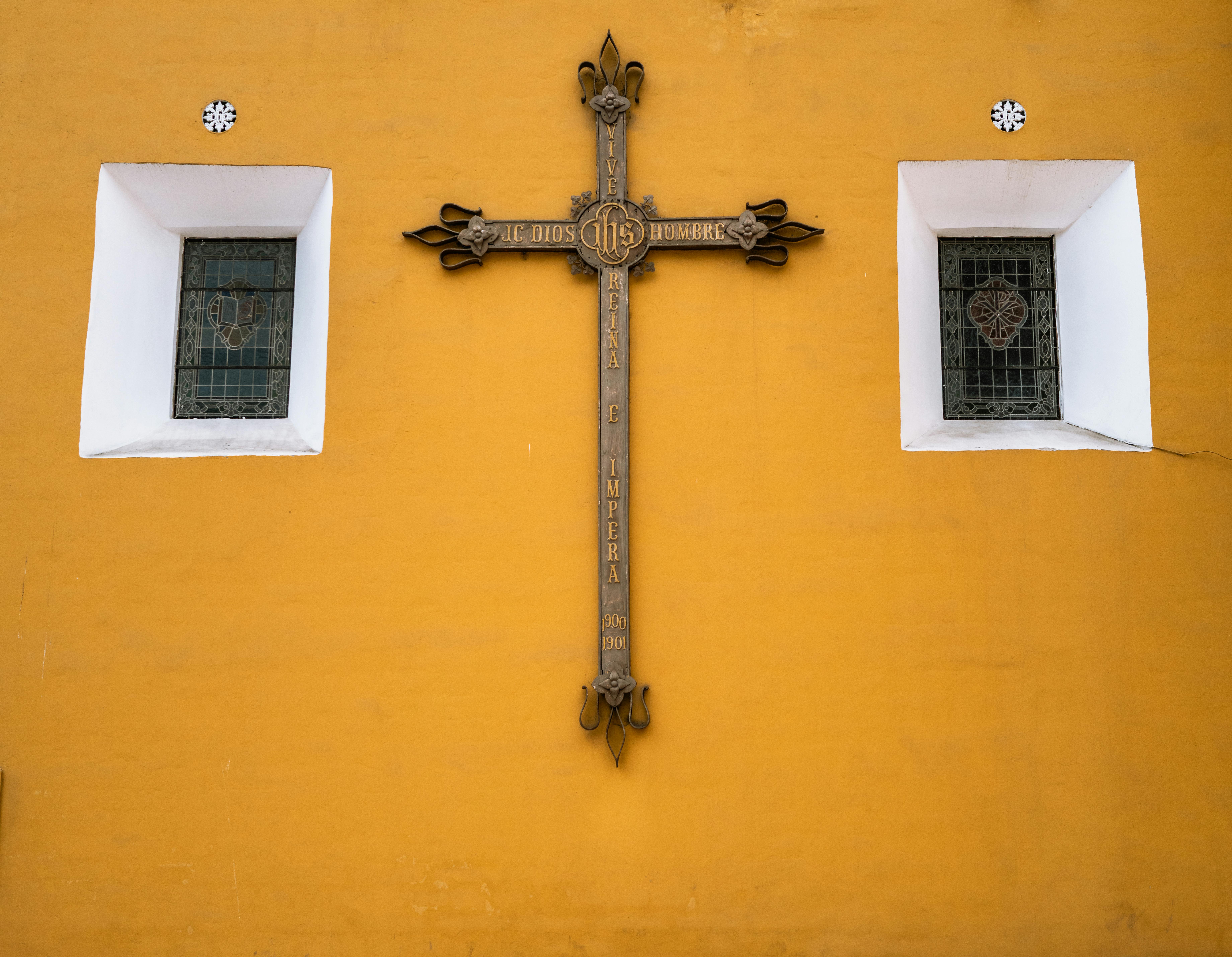 Kostenlos Ein reich verziertes Kreuz mit religiösem Text an einer leuchtend gelben Wand, flankiert von zwei Buntglasfenstern. Stock-Foto