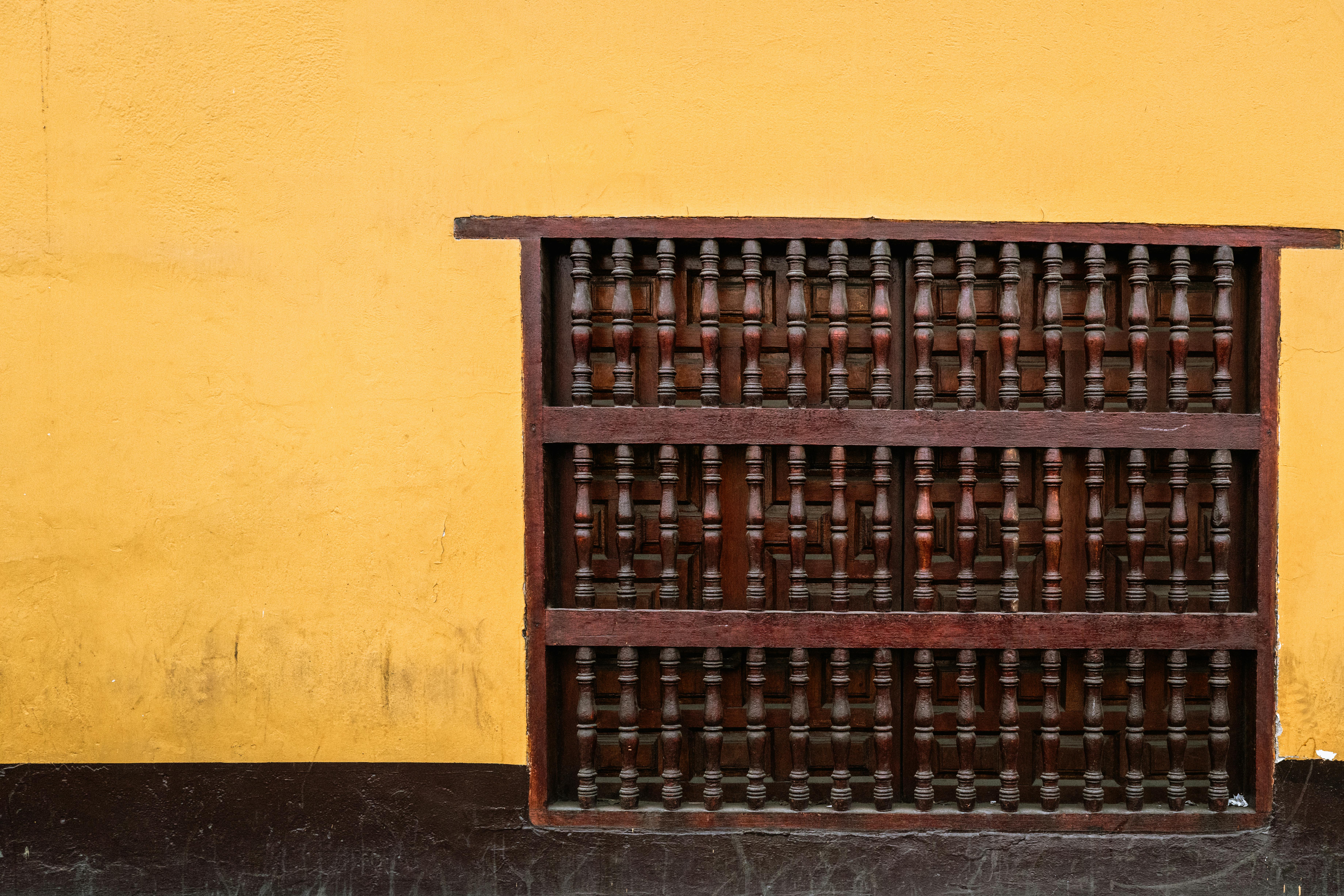 De franc Una finestra de fusta rústica, d'estil gelosia, contra una paret groga vibrant, que mostra l'arquitectura tradicional. Foto d'estoc