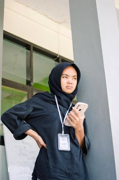 Female in hijab standing with smartphone and ID badge, exuding confidence outdoors.