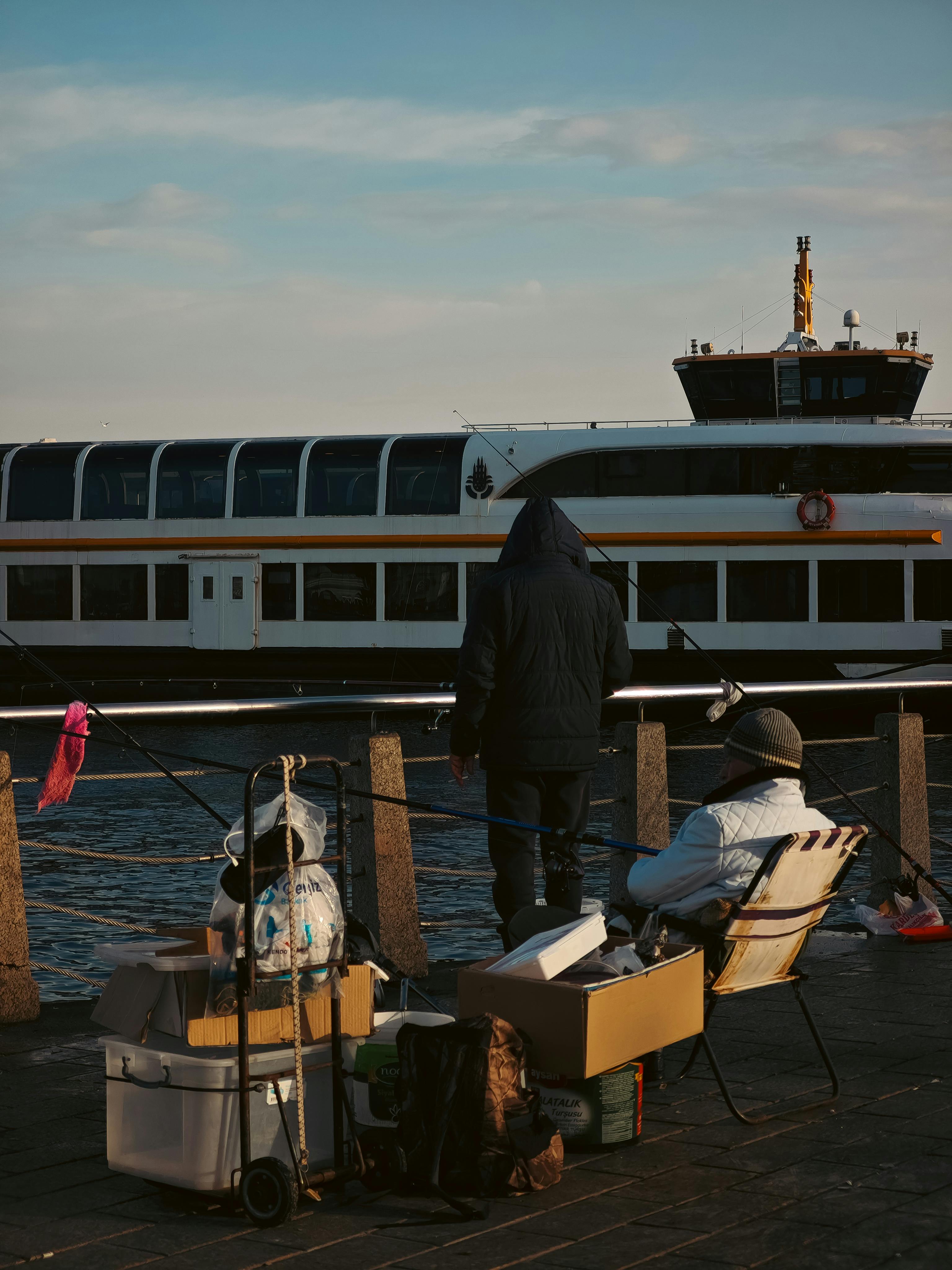 Miễn phí Các ngư dân ngồi bên bến cảng, phía sau là một chiếc phà đang lướt qua, tận hưởng một ngày yên bình. Ảnh lưu trữ