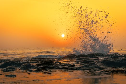 Dramatic sunset over ocean waves crashing against rocks, creating a vivid and warm scene.