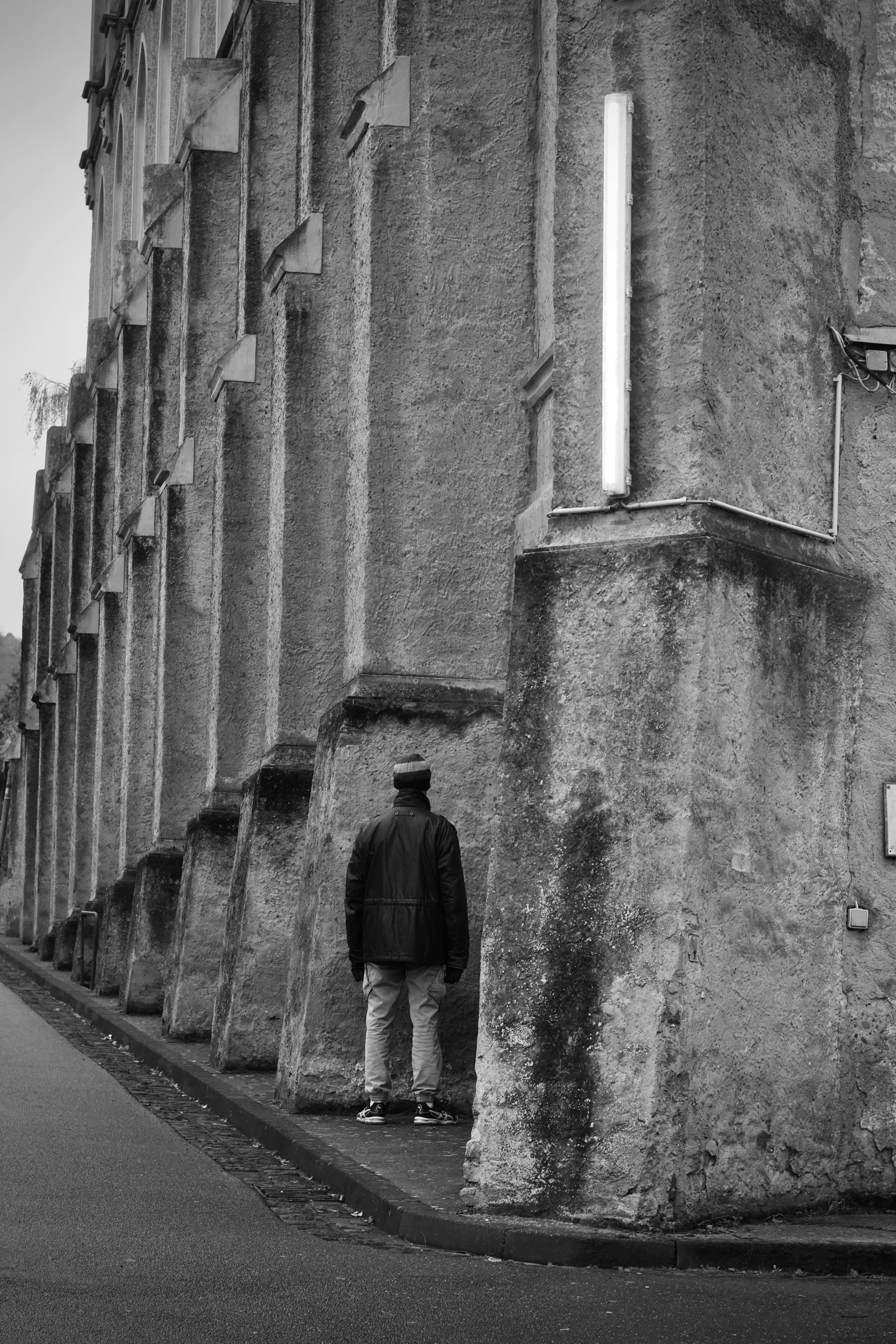 Δωρεάν Μια μοναχική φιγούρα στέκεται δίπλα σε έναν τραχύ πέτρινο τοίχο γοτθικού ρυθμού, αποτυπωμένη σε μονόχρωμη μορφή για δραματικό αποτέλεσμα. Φωτογραφία από στοκ φωτογραφιών