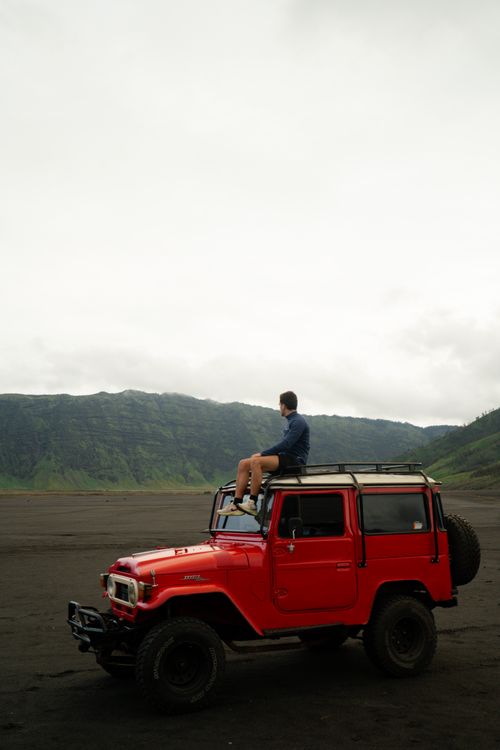 Free Man enjoying the view from a red jeep on a barren landscape with green hills in the background. Stock Photo