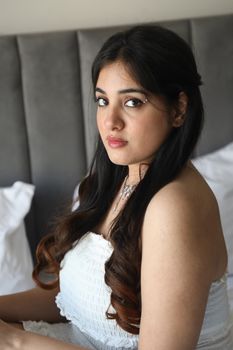 A young woman in a white dress sitting on a bed, captured in natural light.