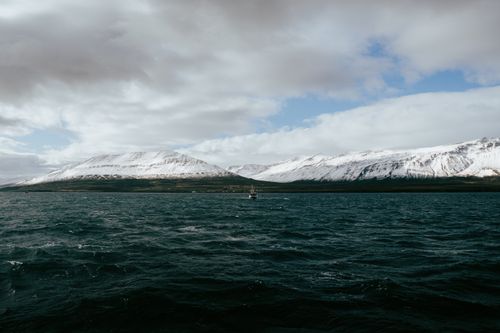 Free A sailboat glides through a fjord in Árskógssandur, Iceland, with snow-covered mountains in the background. Stock Photo