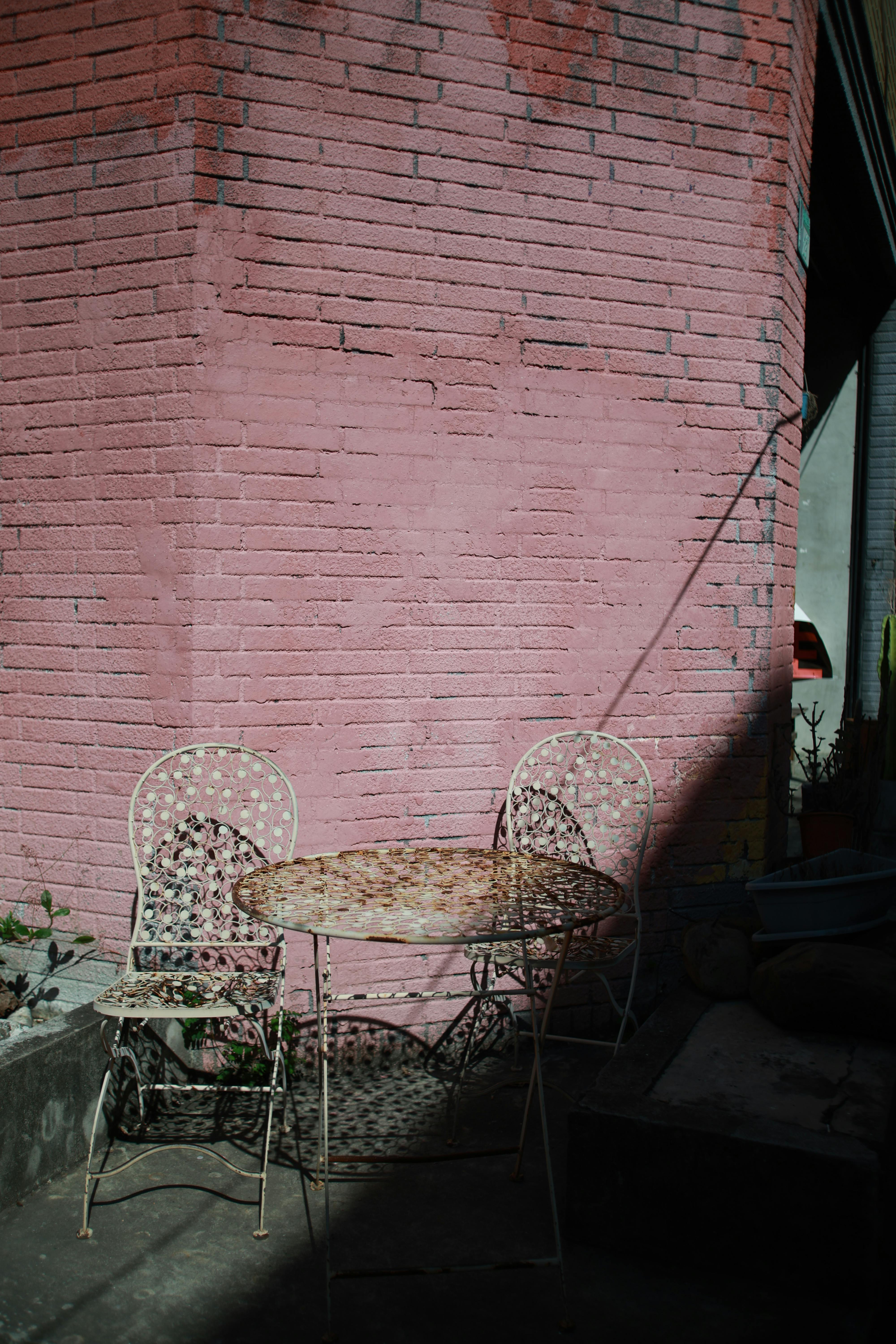 bezplatná Útulné venkovní posezení s vintage kovovými židlemi a stolem u růžové cihlové zdi, ideální pro slunečný den. Základová fotografie