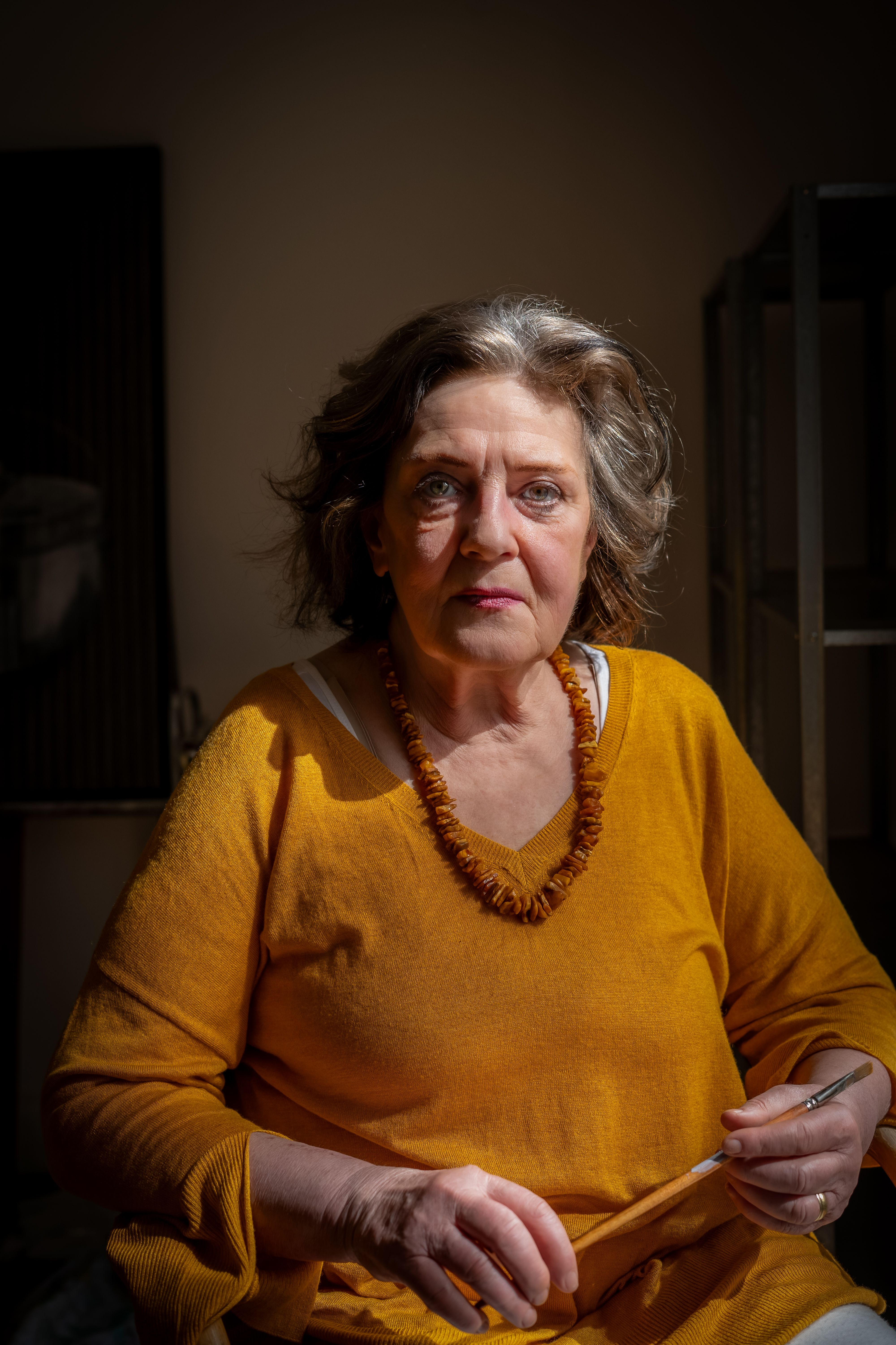 Elderly woman in vibrant clothing sits thoughtfully indoors, bathed in light.