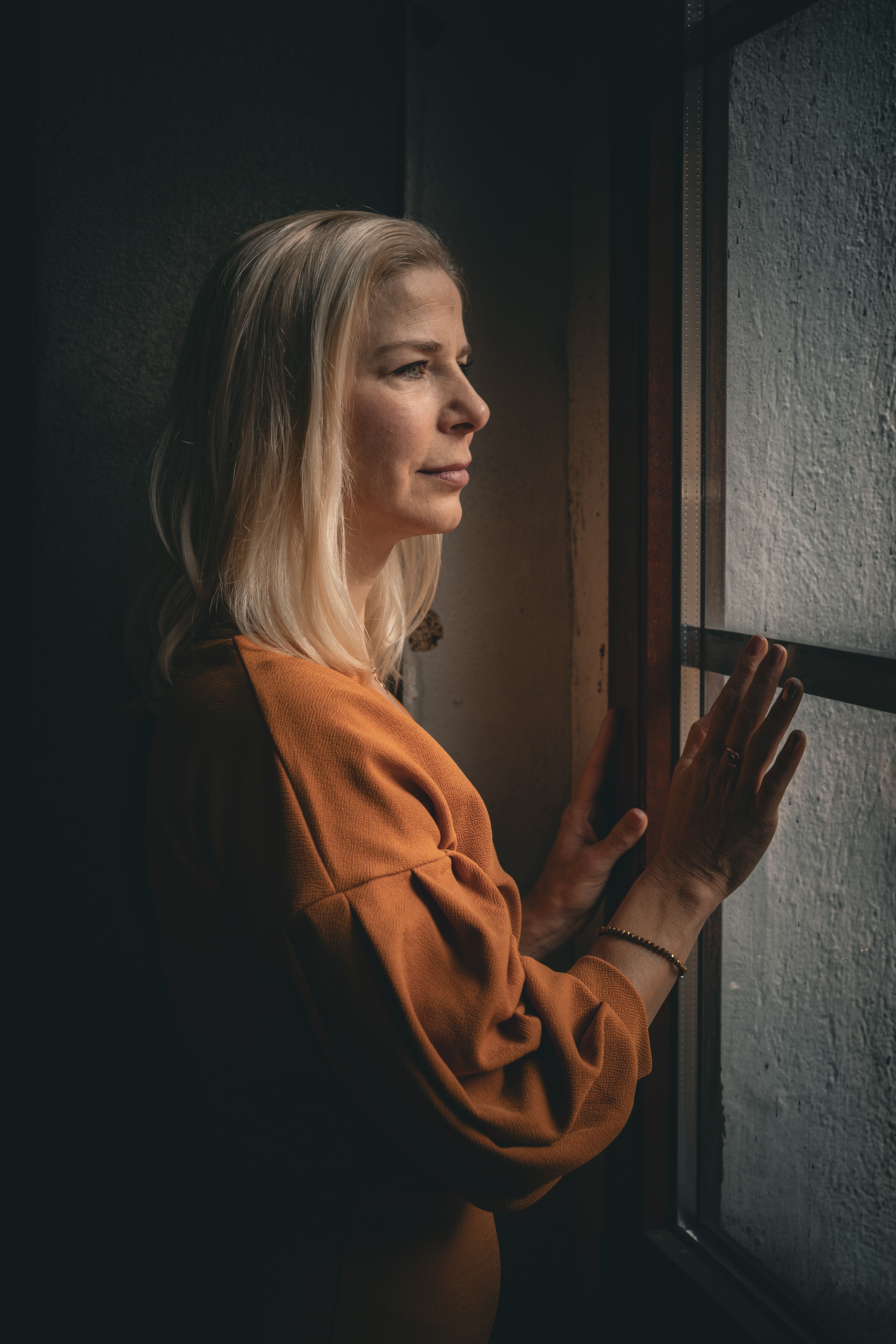 A contemplative woman stands by a window, bathed in warm, soft light, indoors.