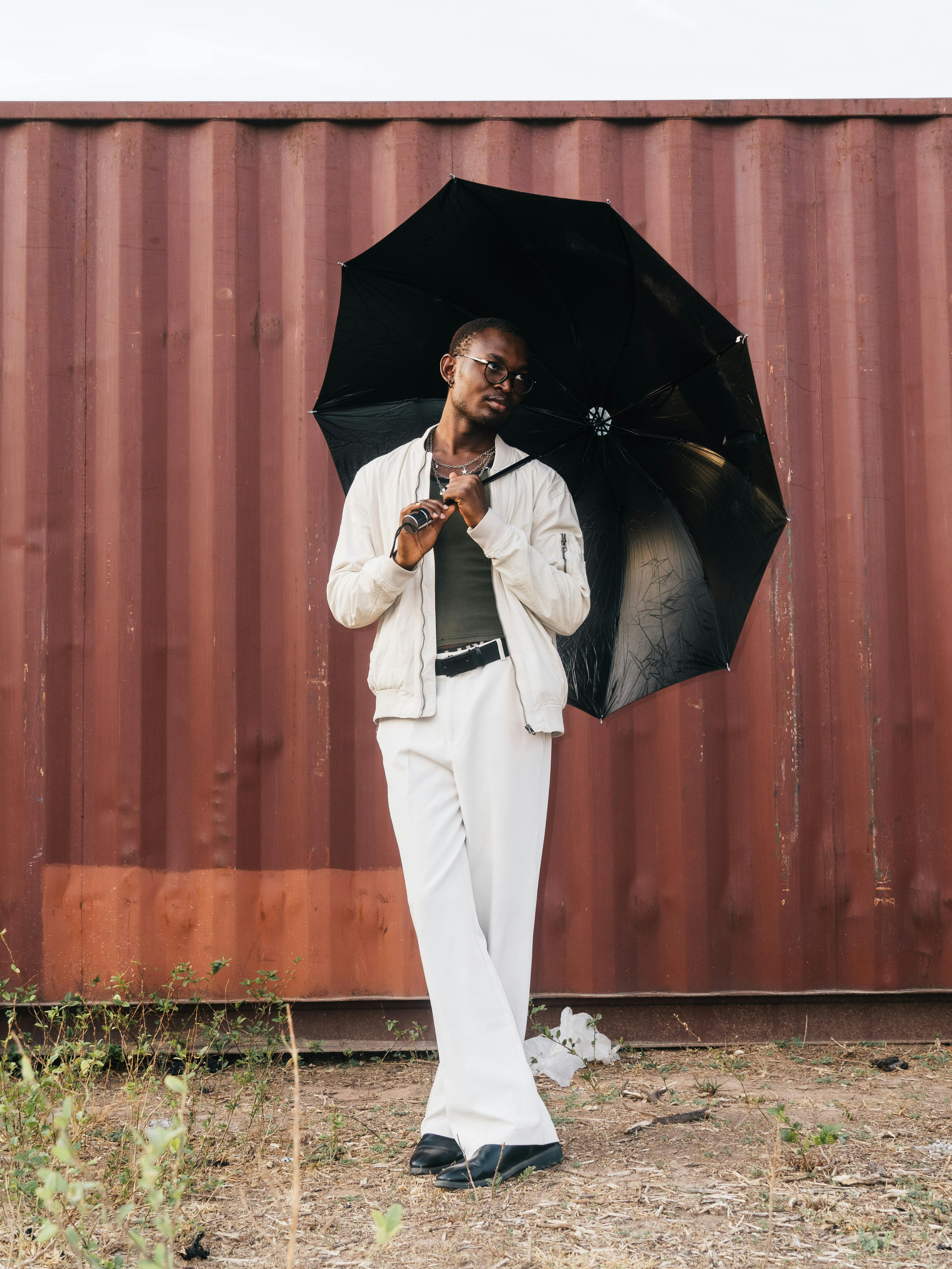 Gratis En moderigtigt klædt person, der holder en sort paraply, poserer stilfuldt op mod en container-baggrund. Lagerfoto